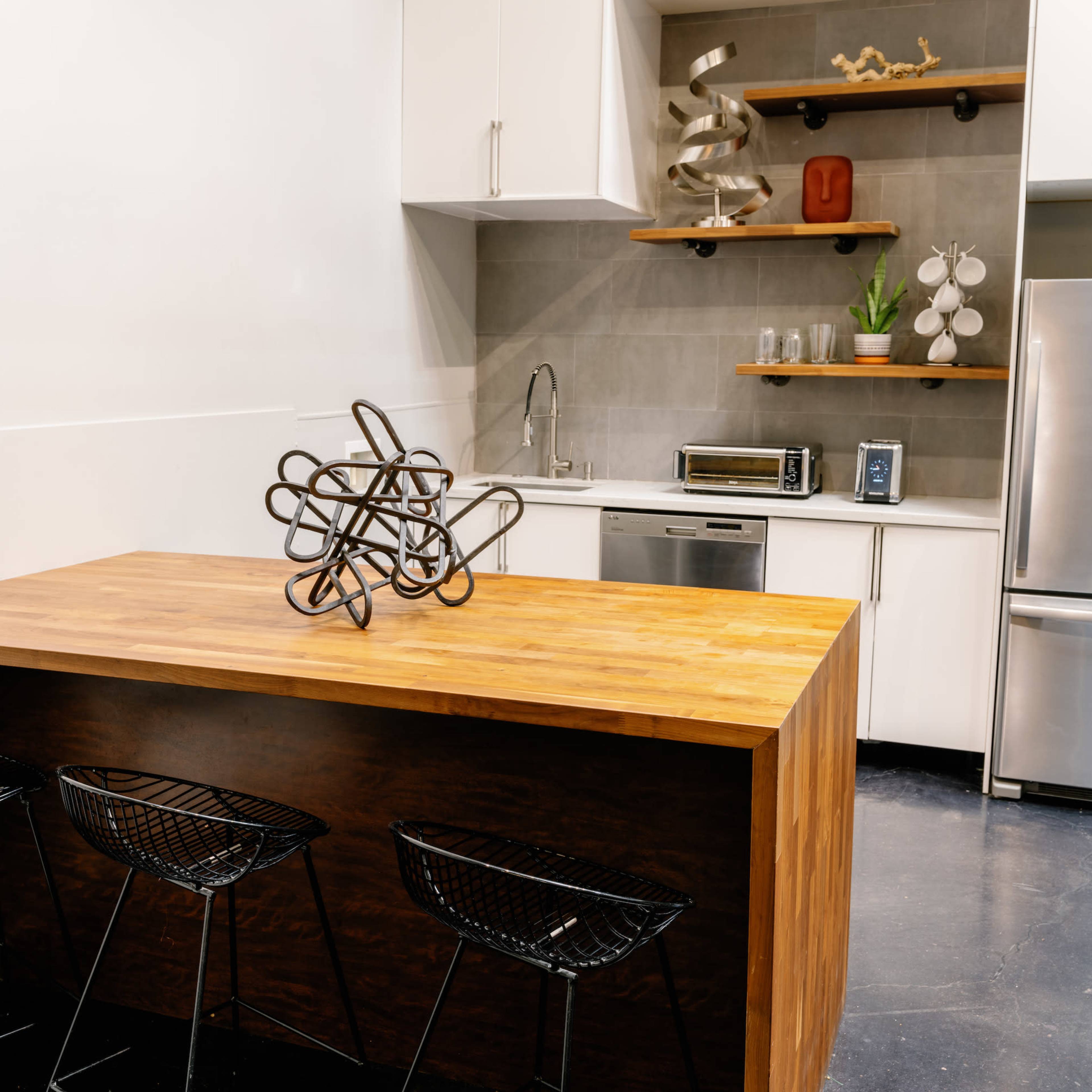 A modern kitchen features a wooden island with three wire bar stools, surrounded by stainless steel appliances and open shelving.