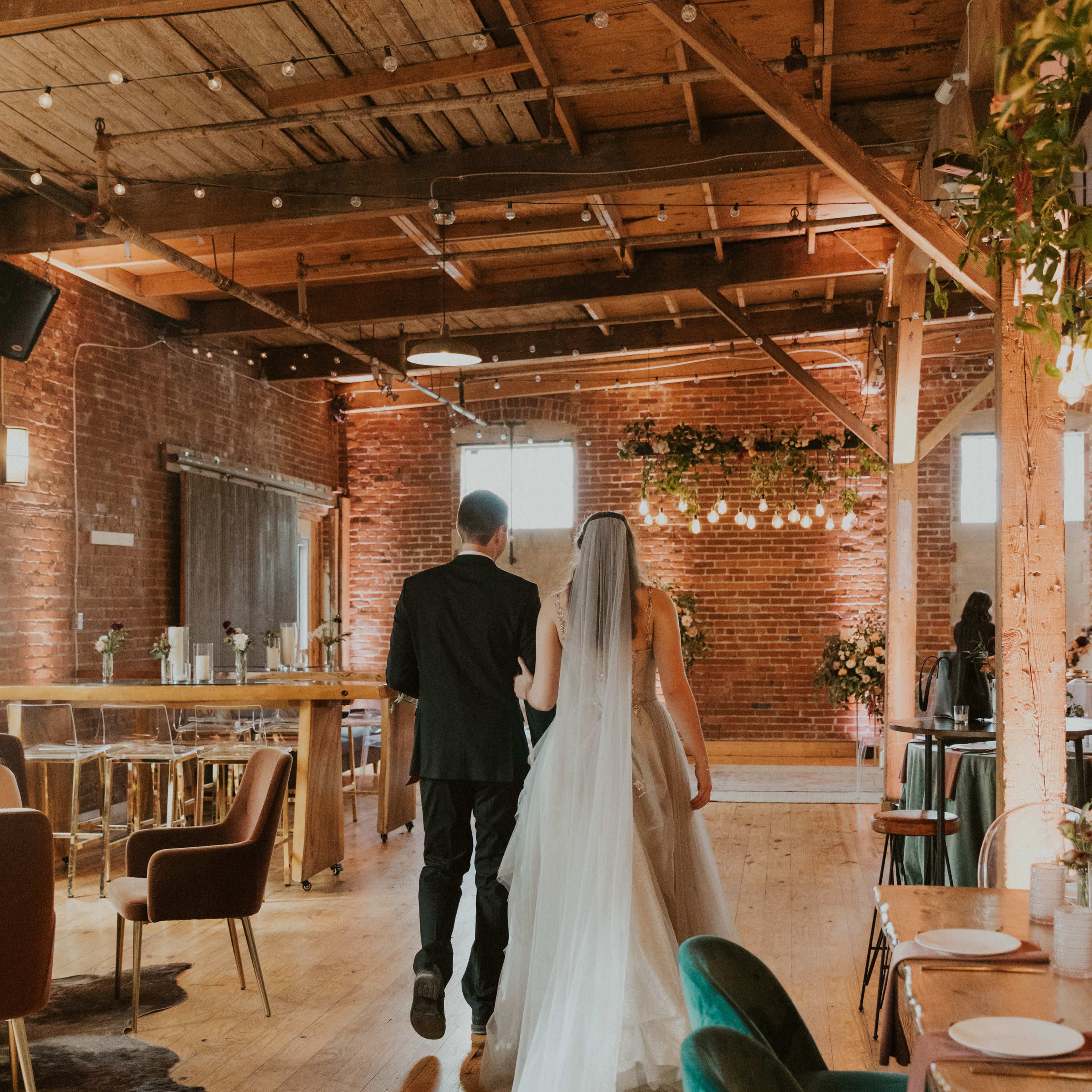 A couple walks through a rustic event space adorned with wooden beams and ambient lighting, with the bride wearing a flowing veil.