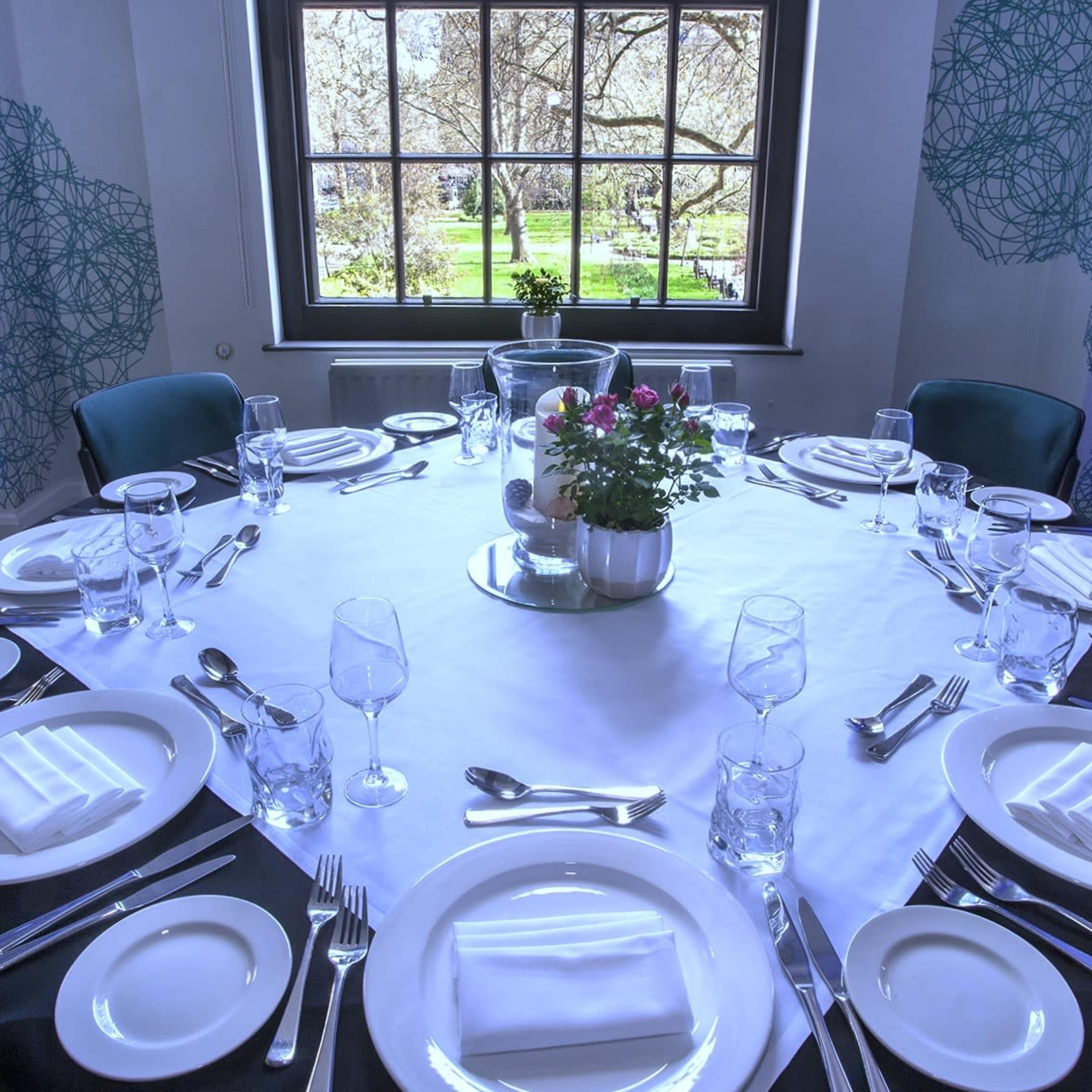 A round dining table is set for multiple guests, featuring white plates, glasses, and a central floral arrangement, with a view of a garden visible through a large window.