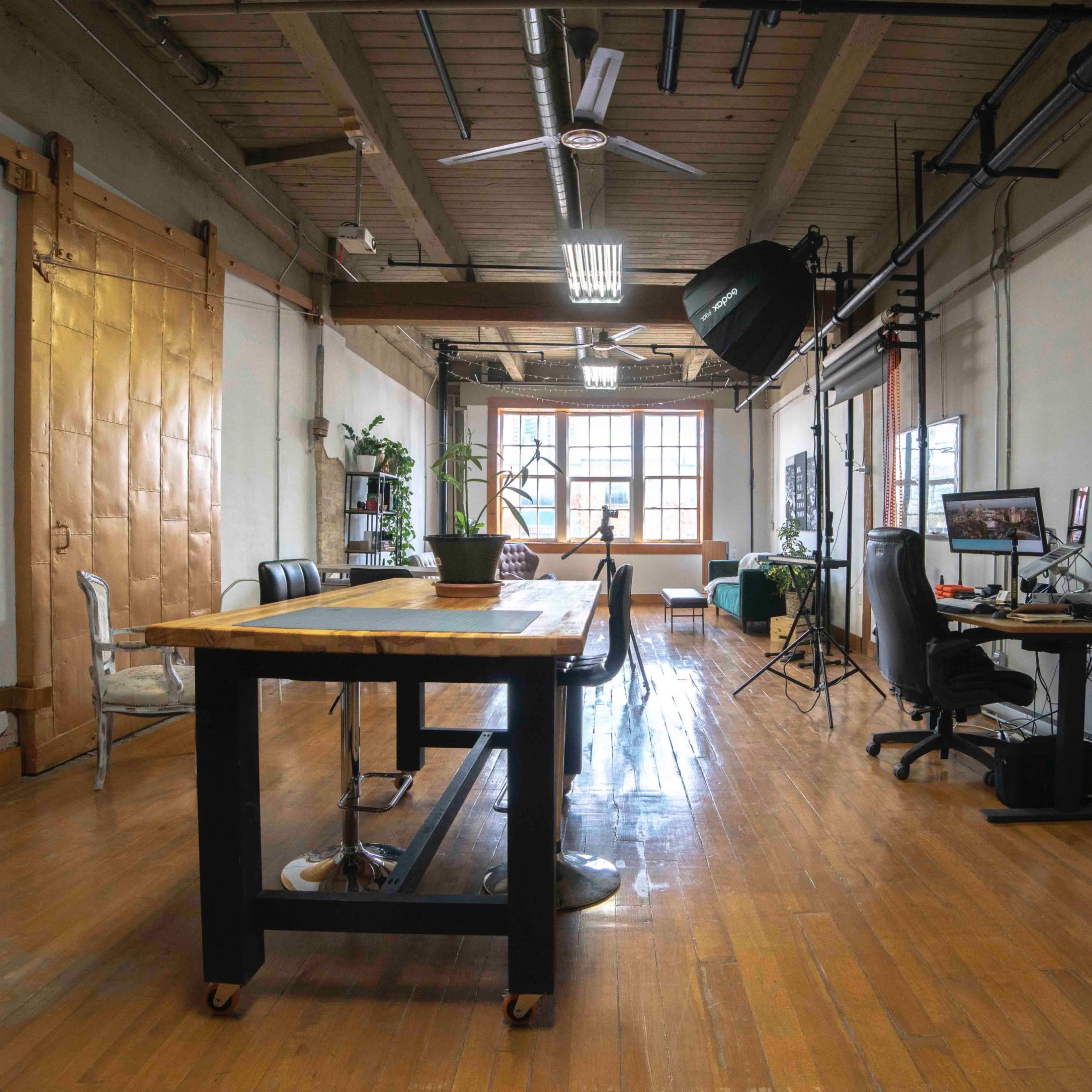 A spacious room with large windows, wooden flooring, a long table, chairs, and various equipment along the walls.