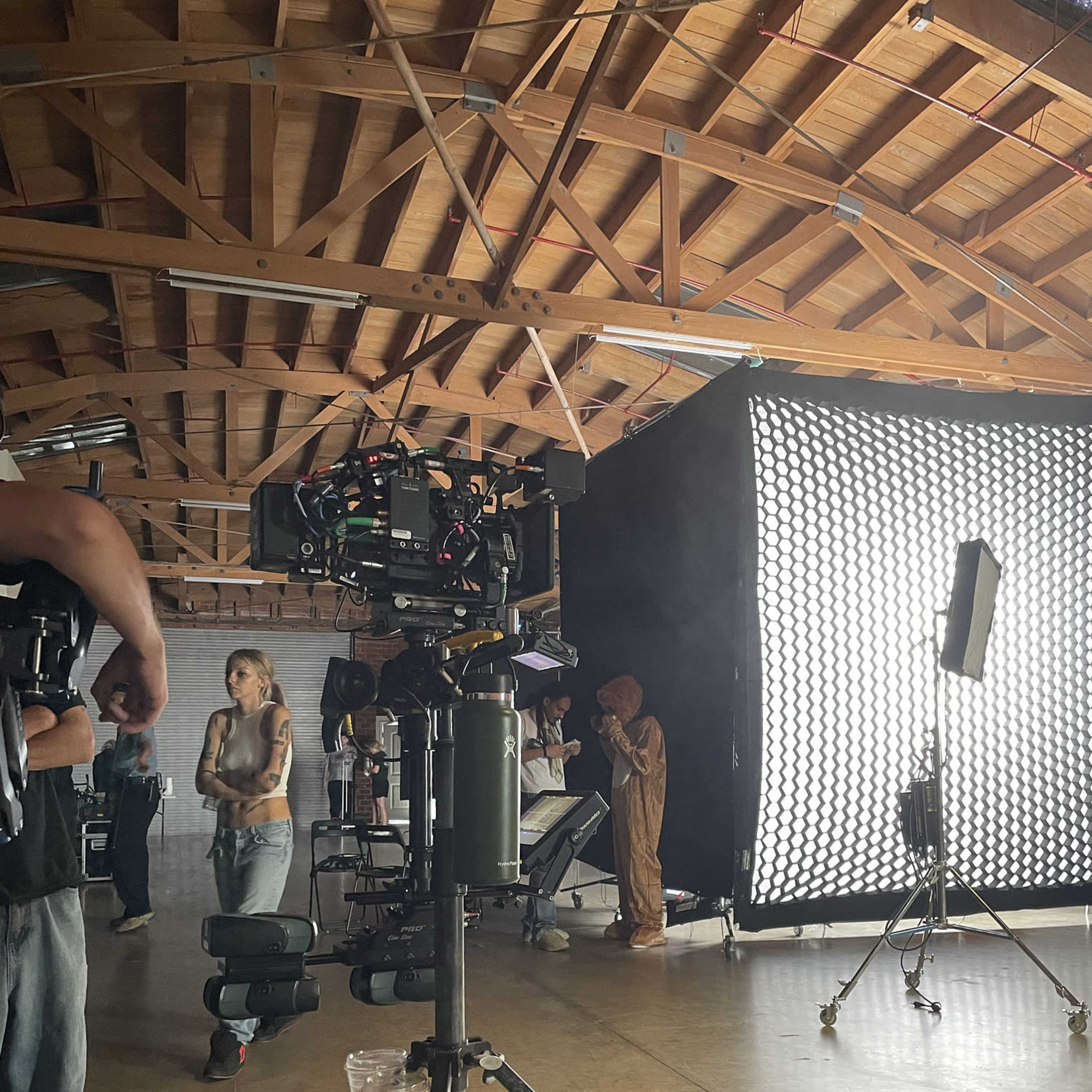 A film crew sets up equipment in a spacious studio, featuring cameras, lighting, and a figure in a costume behind a large softbox.