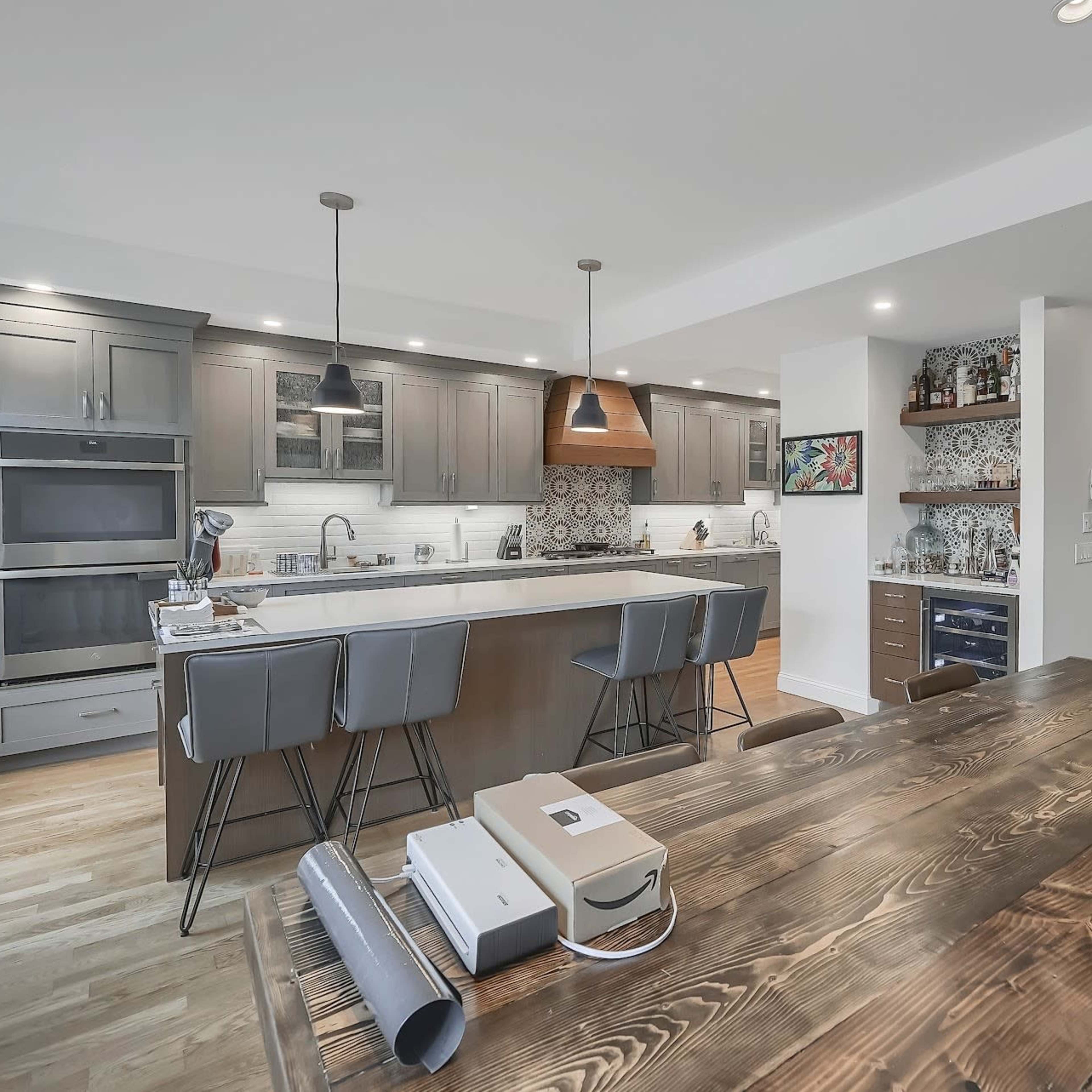 A modern kitchen features a large island with bar stools, wooden dining table, and stylish light fixtures.