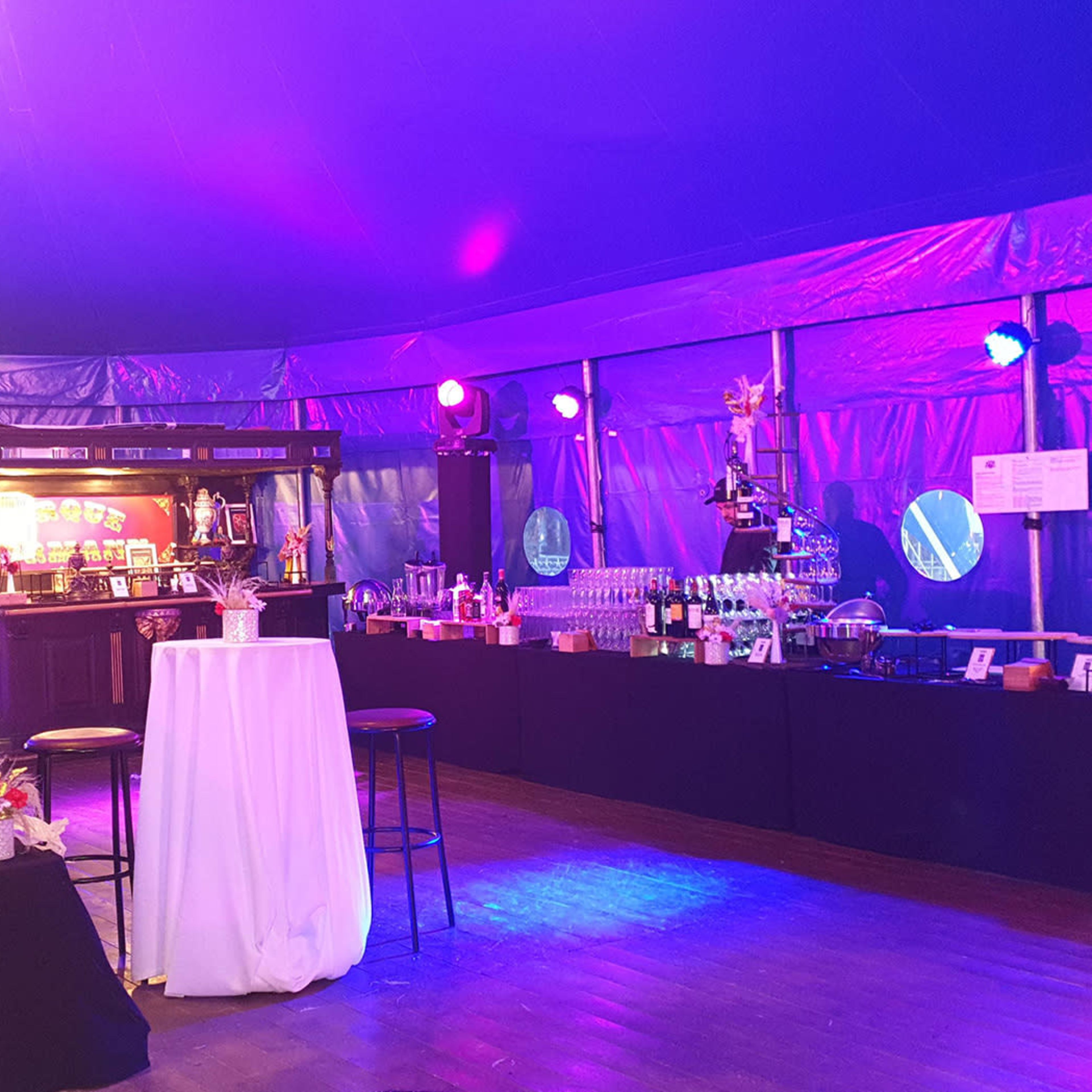 A brightly lit indoor event space with a bar counter, several high tables, and colorful lighting under a tent.