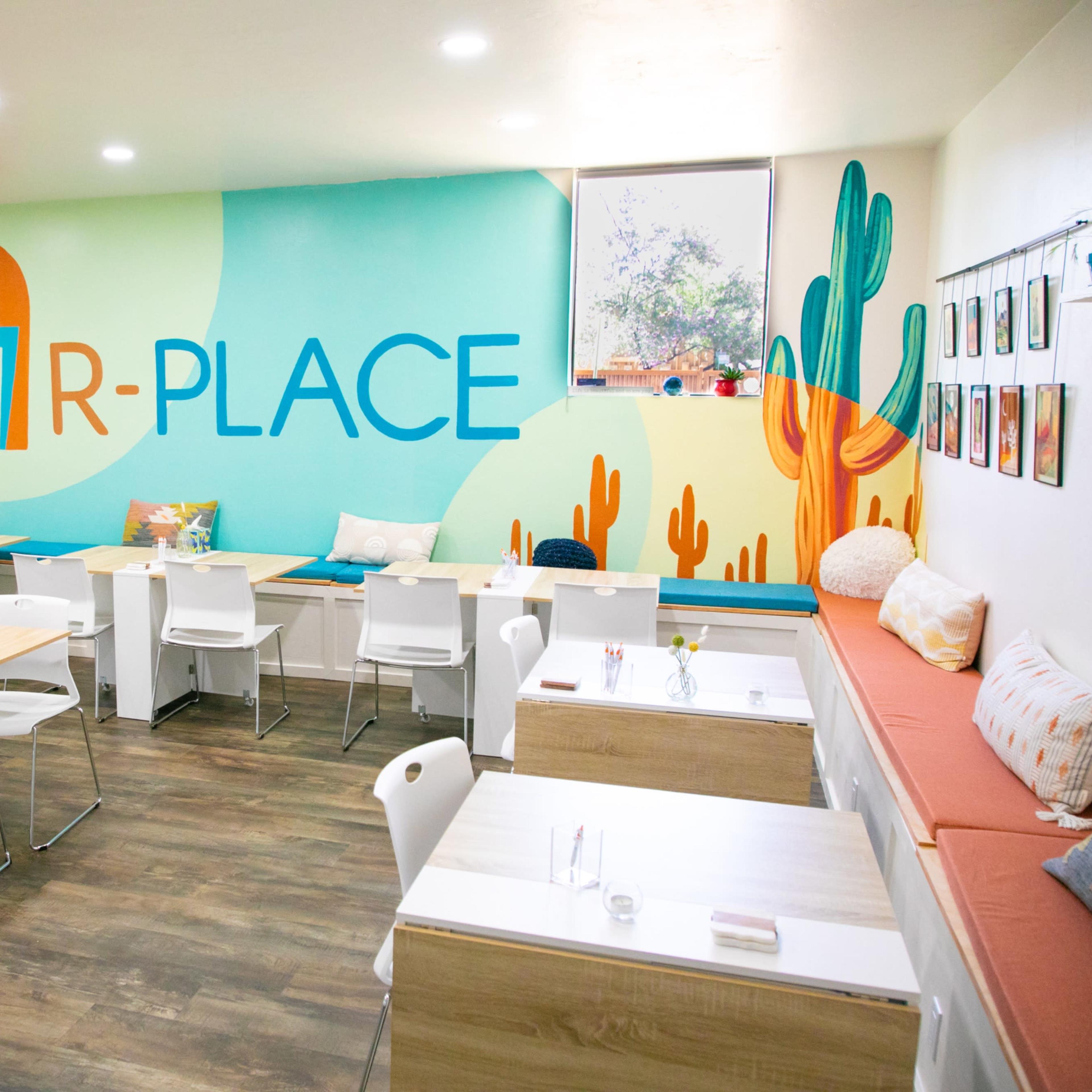 A brightly colored café interior featuring a large mural with the word "R-PLACE," surrounded by cacti decor, tables, and seating areas.