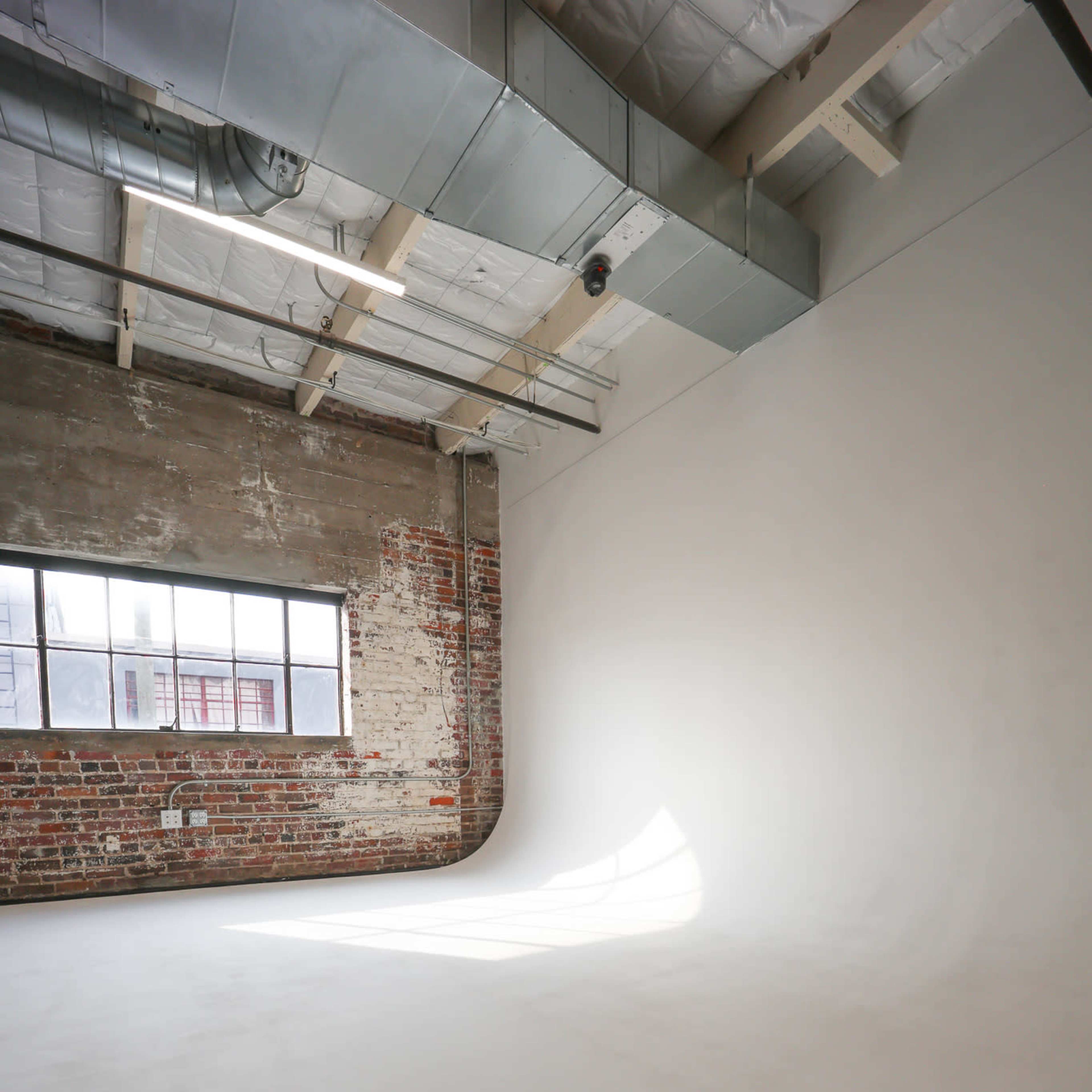 An empty studio space with a smooth white floor, exposed brick walls, and a large window letting in natural light.