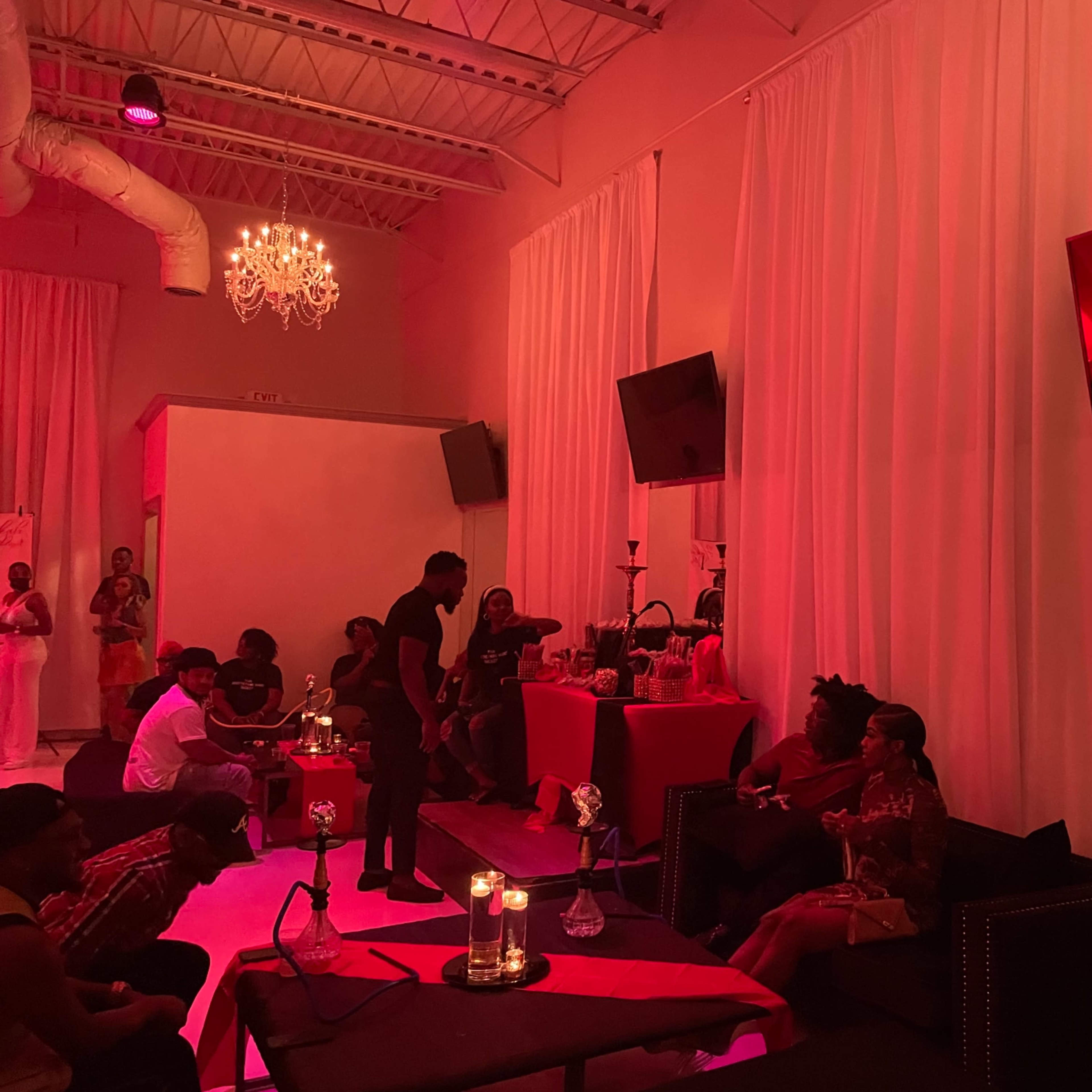 A dimly lit event space with red lighting, featuring guests seated around tables with candles and decorative items.