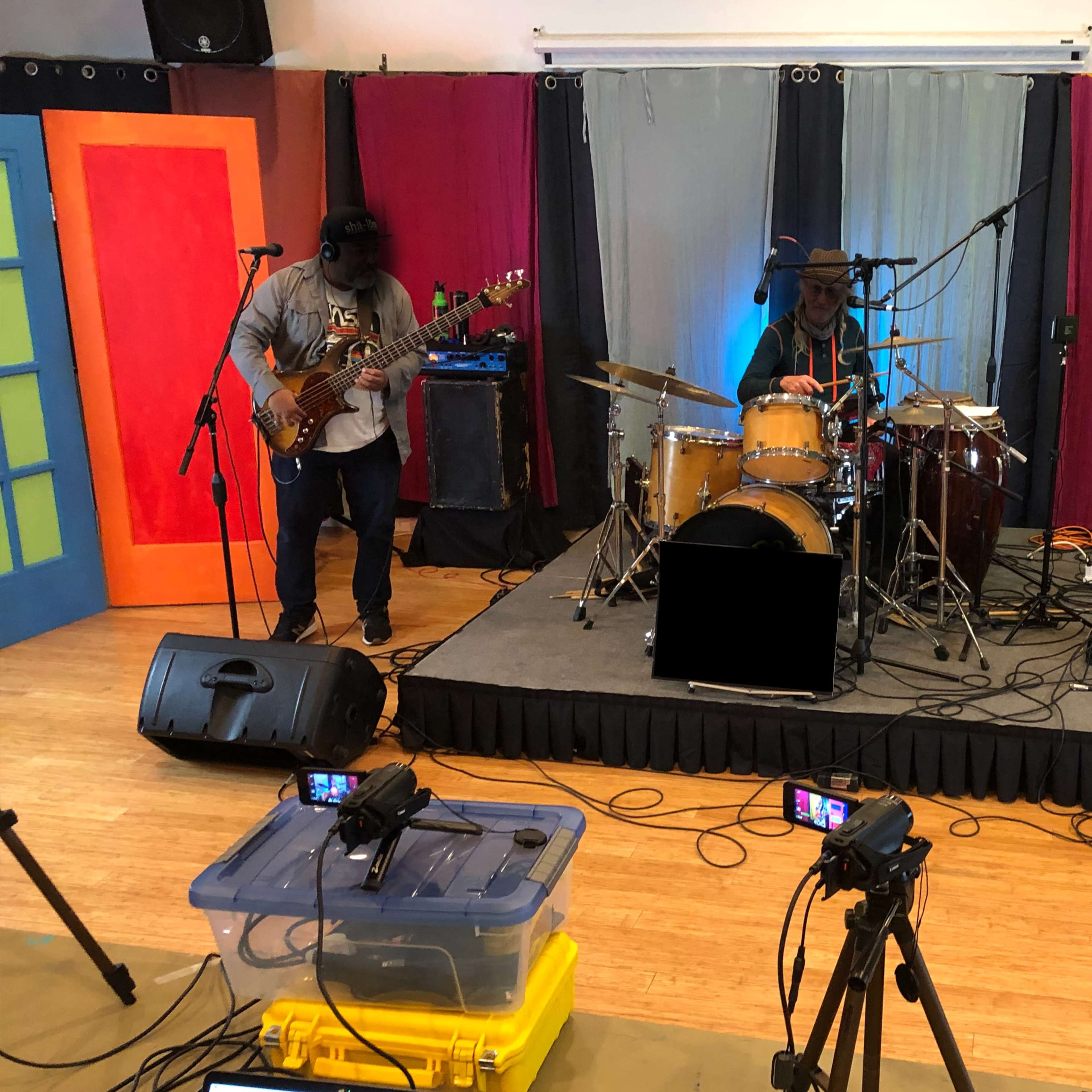 Three musicians perform on a small stage featuring drums, a bass guitar, and an electric guitar, surrounded by colorful backdrops and audio equipment.