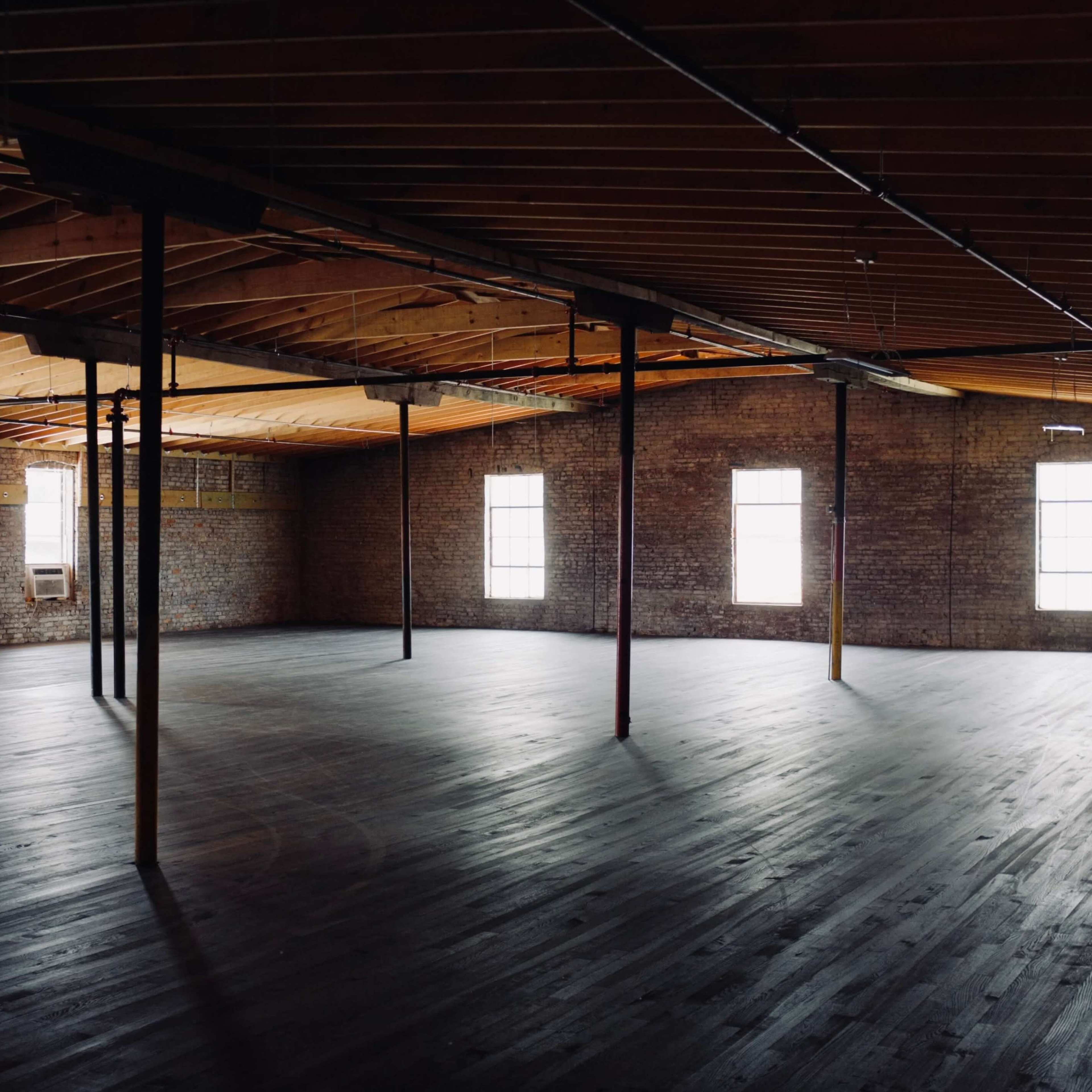 An empty, industrial-style room features exposed brick walls, wooden beams overhead, and large windows allowing natural light to enter.