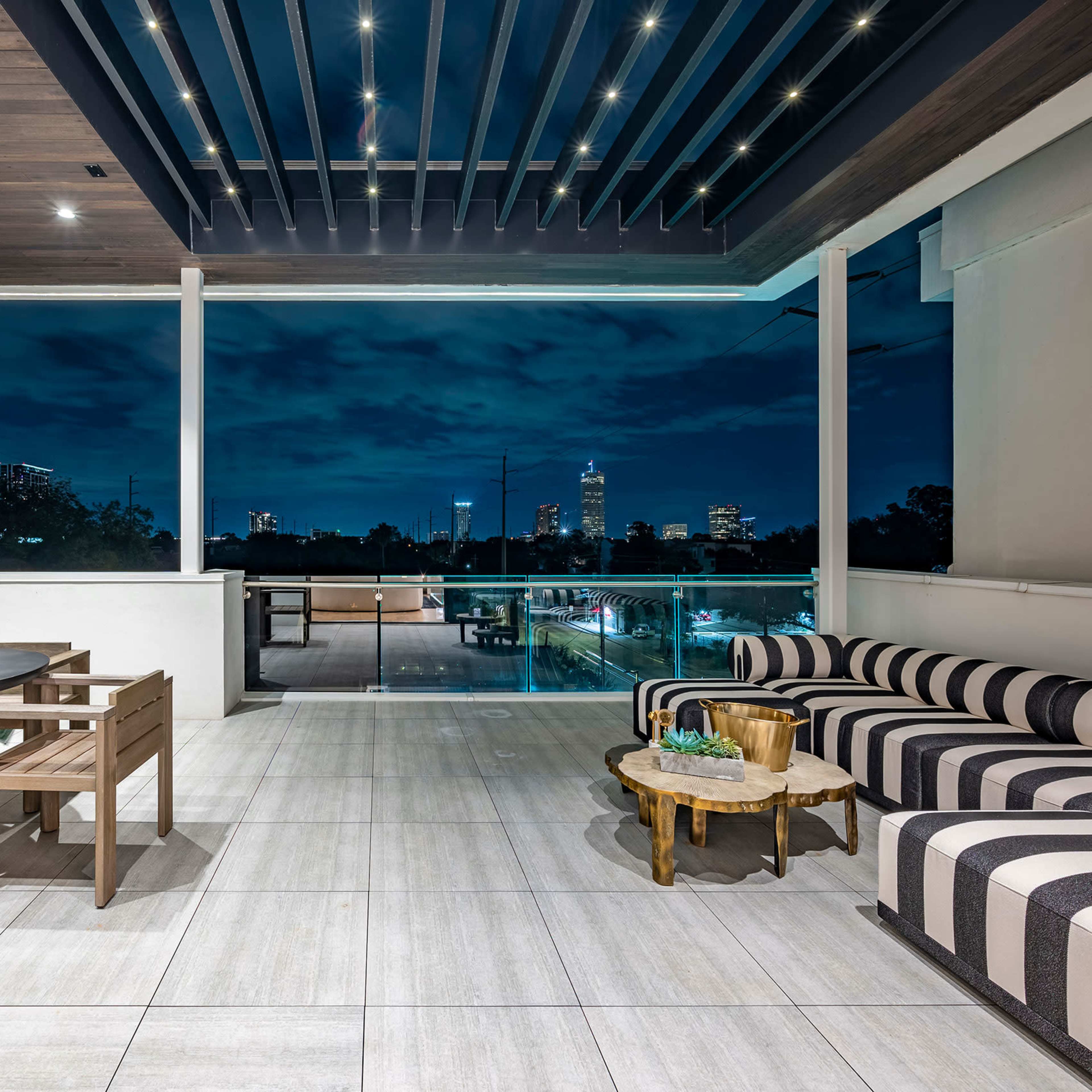 A modern outdoor patio with wooden seating, a dining set, a grill, and city skyline views at dusk.