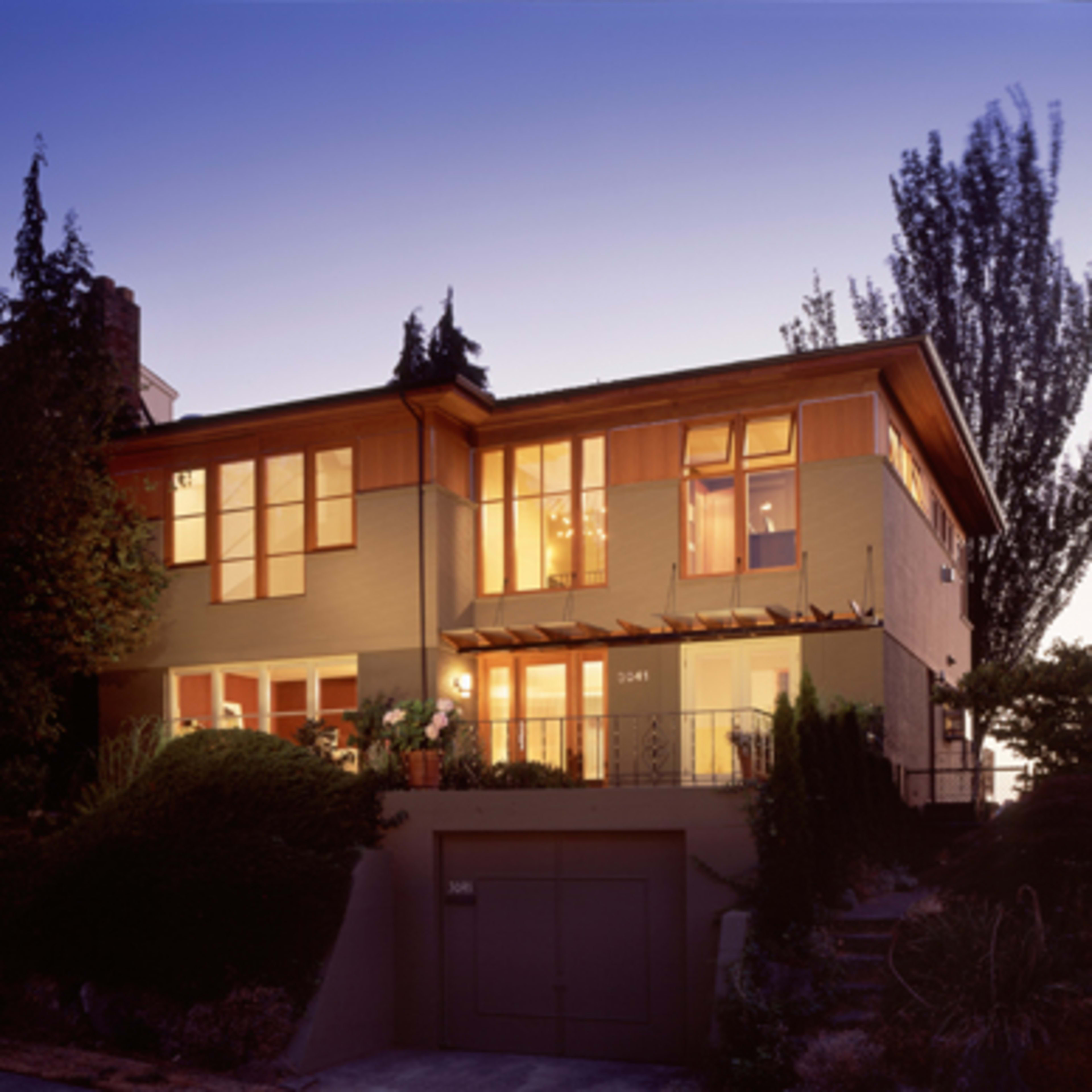 A modern two-story house with large windows and a landscaped front garden is illuminated at dusk.