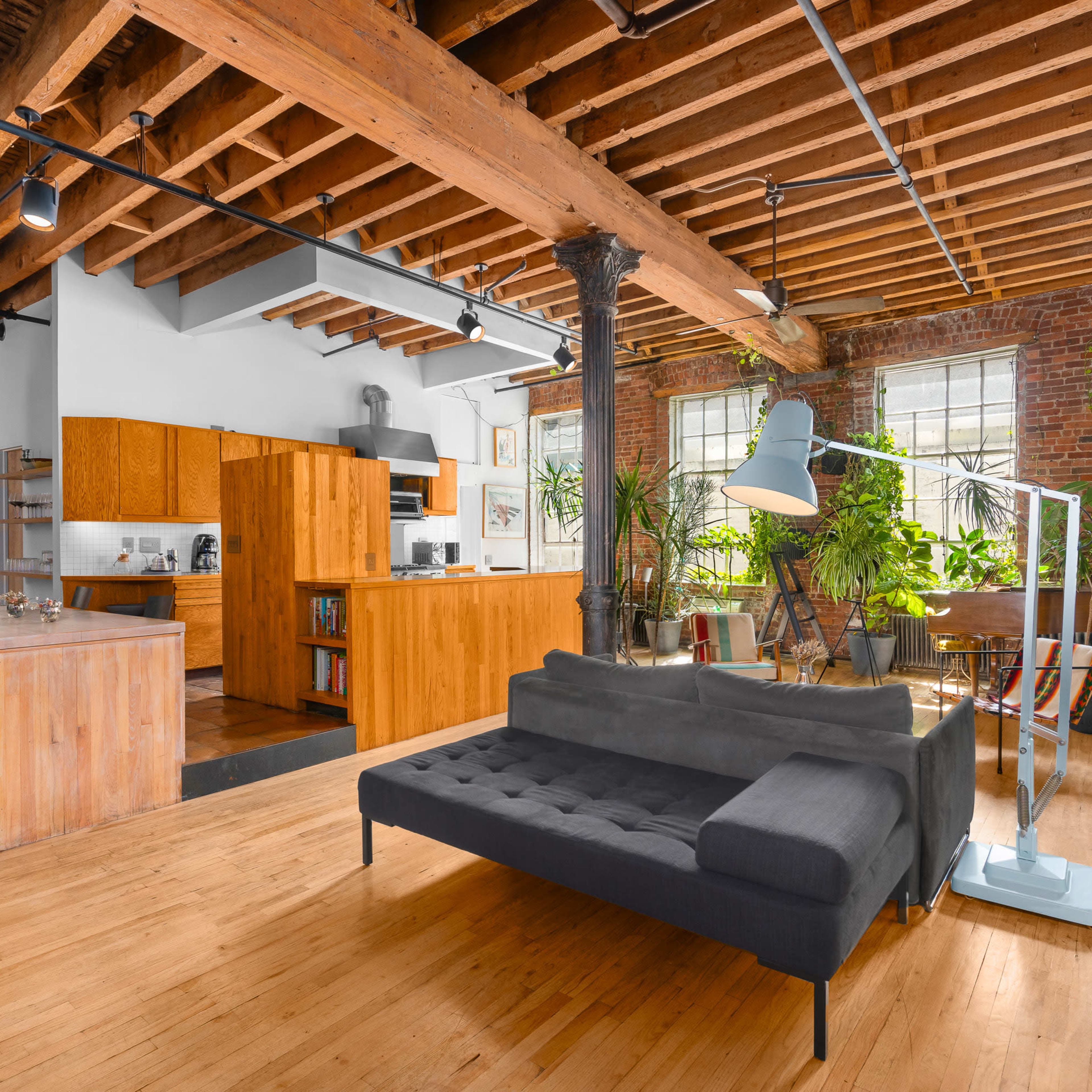 A spacious loft-style living area with exposed wooden beams, modern furniture, and large windows filled with plants.