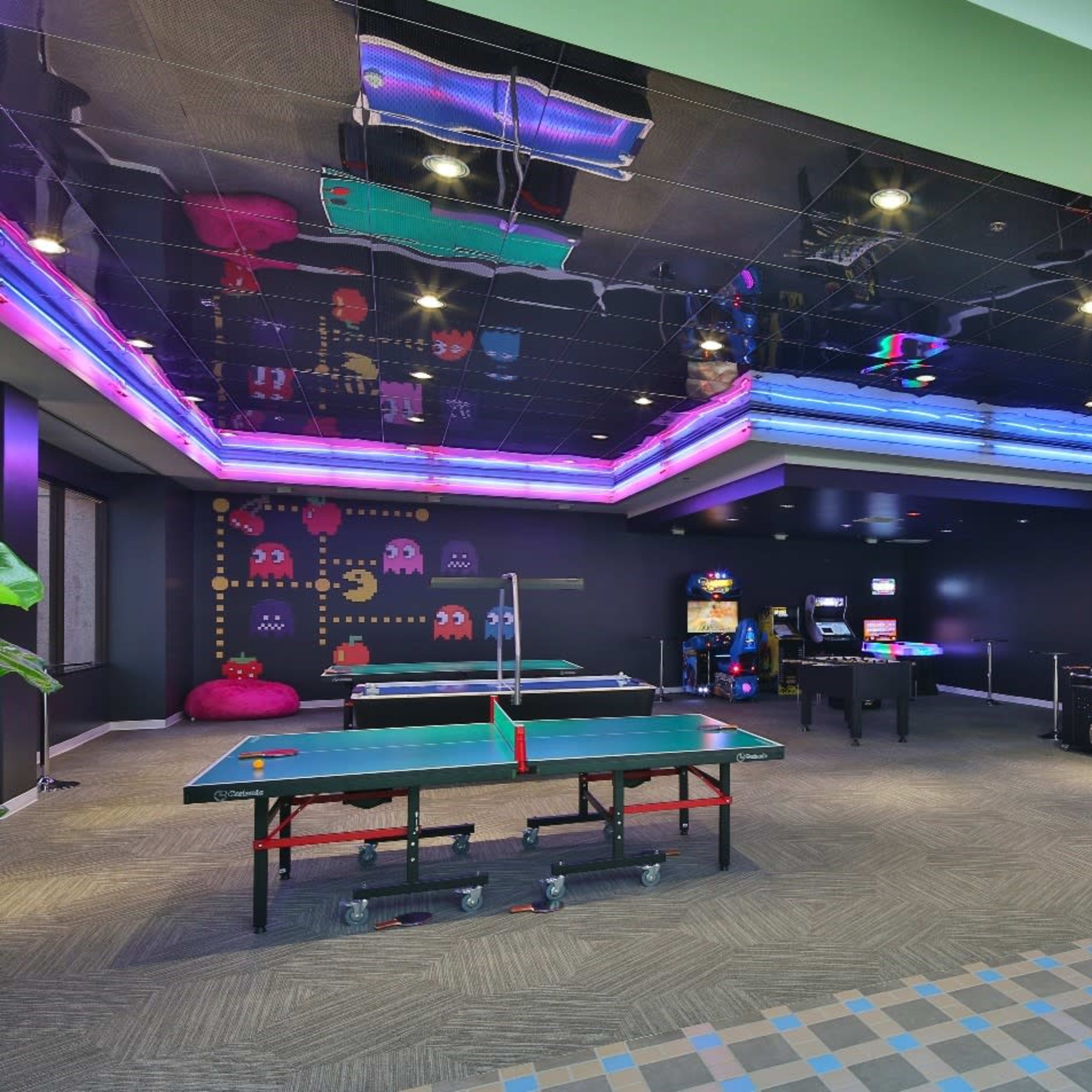 A game room featuring a ping pong table, arcade machines, and vibrant neon lights on the ceiling.