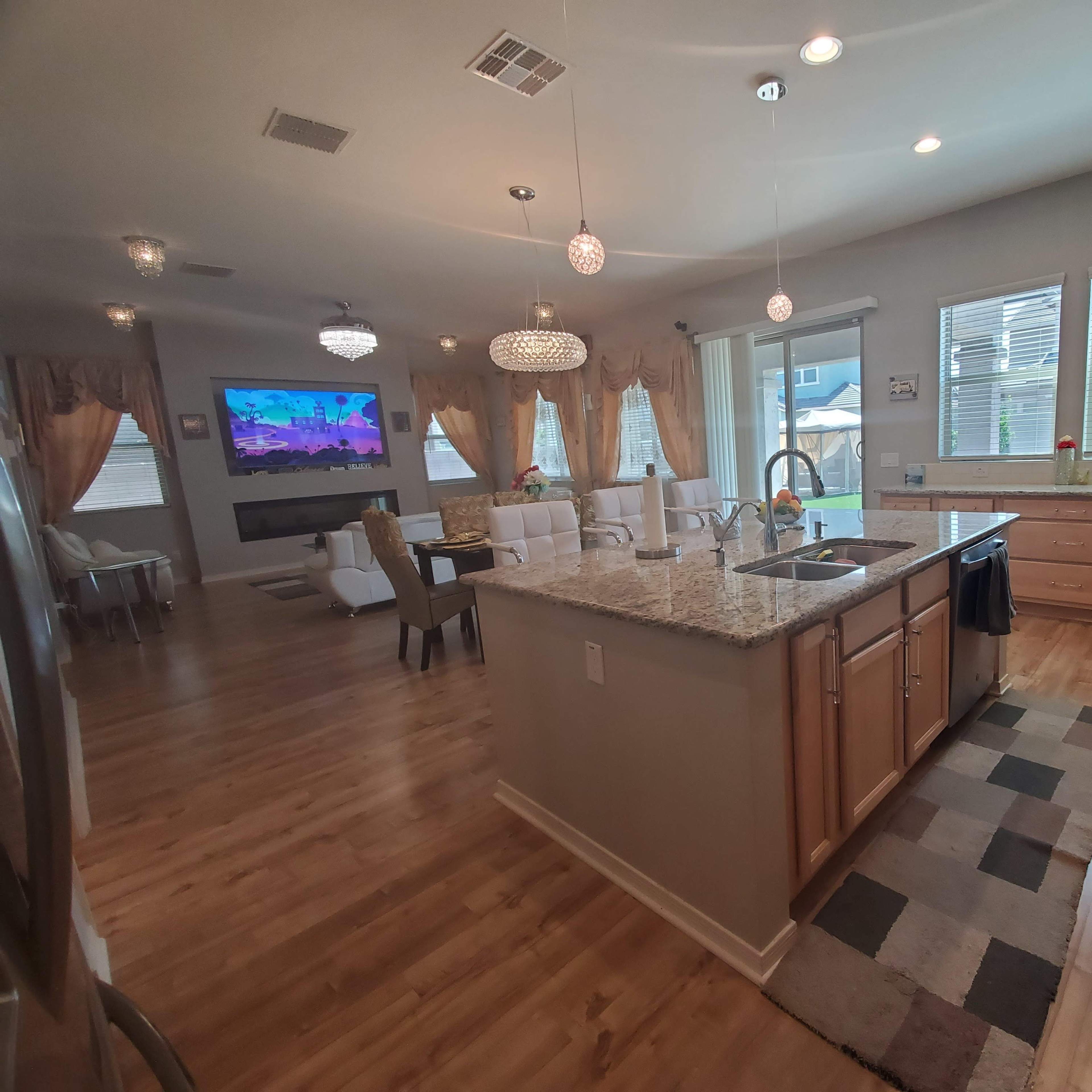 A spacious kitchen with wooden cabinets, a central island, and a living area in the background featuring a large television and light-colored furniture.