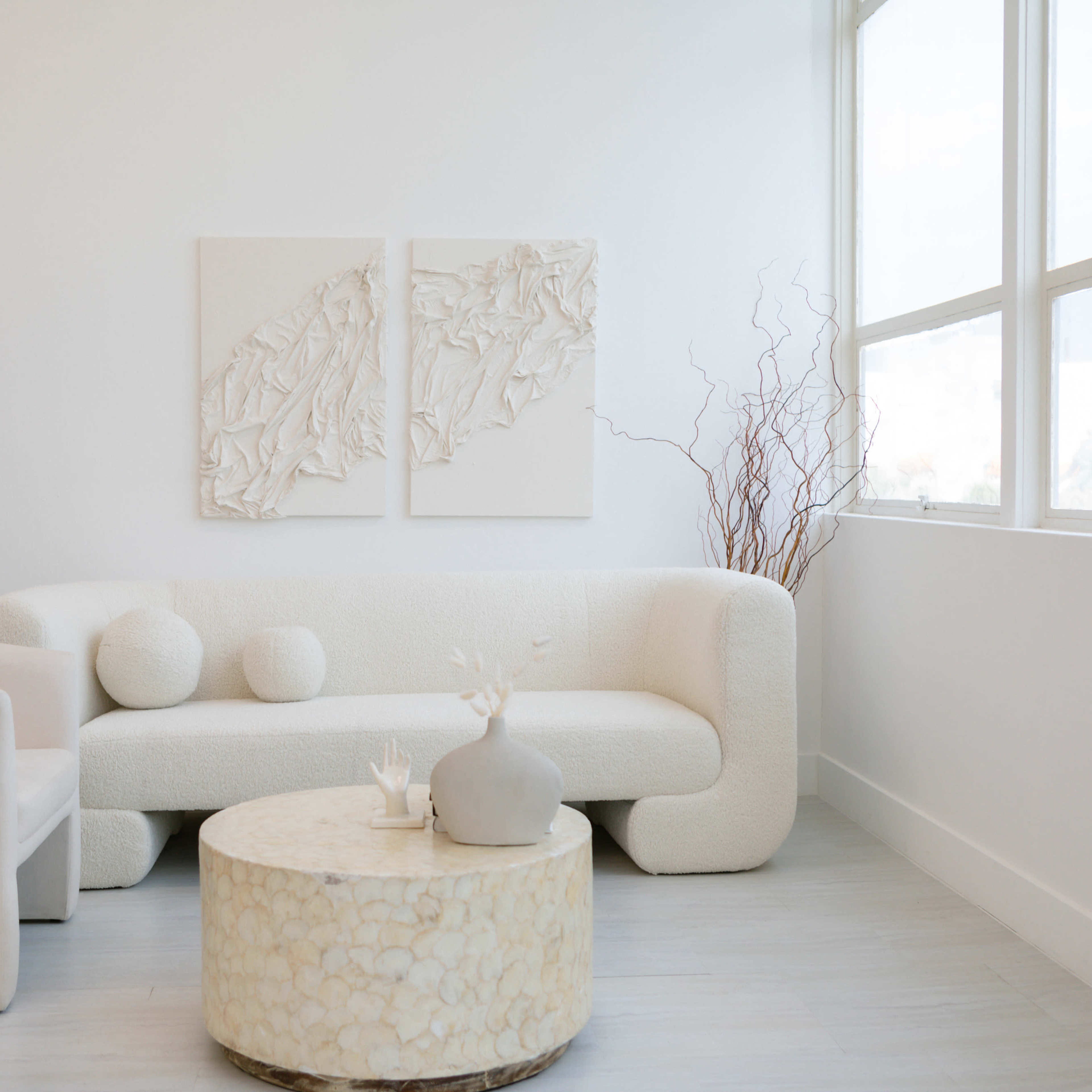 A minimalist living room with a white curved sofa, a round coffee table, and two textured wall artworks, illuminated by natural light from large windows.