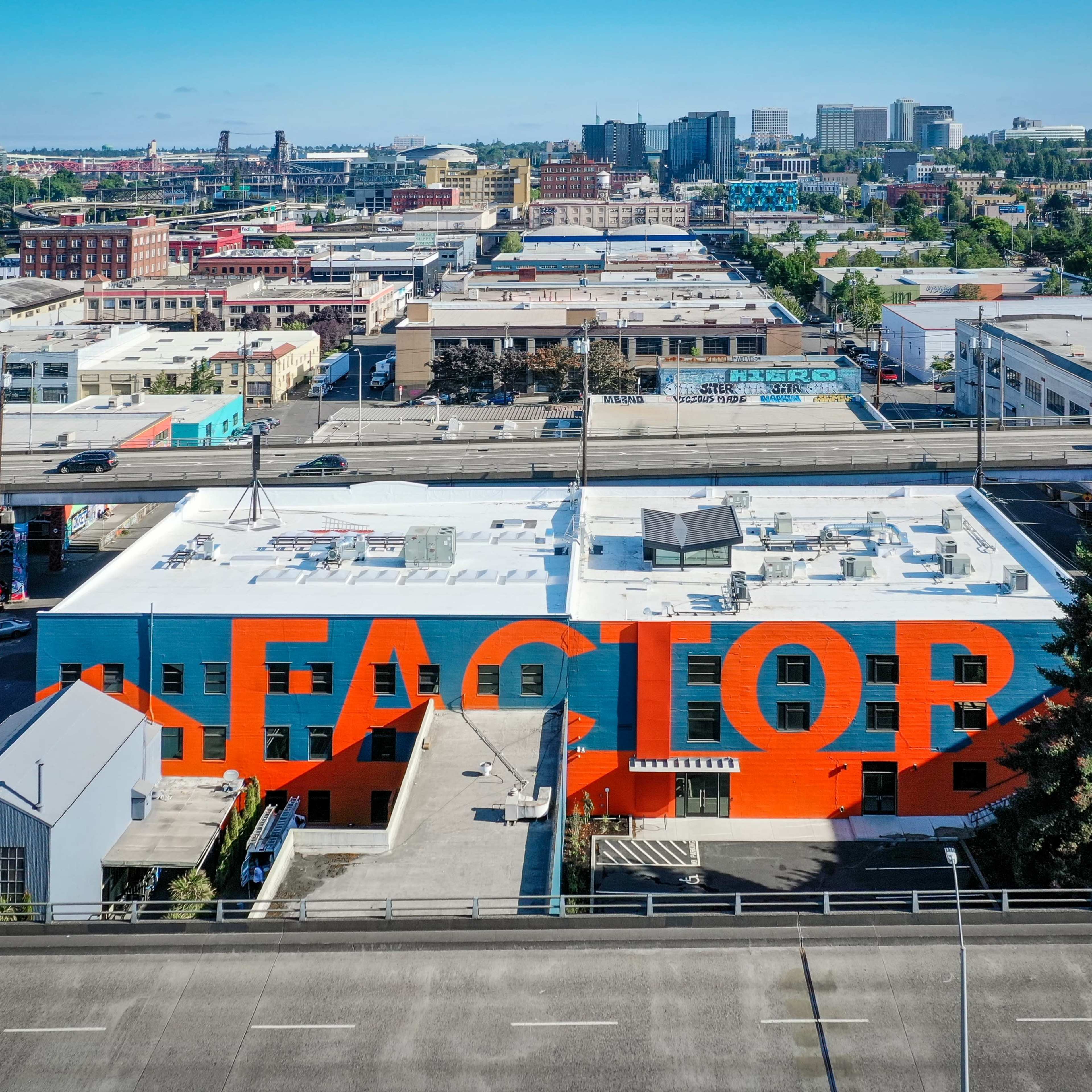 An industrial building with large, bold lettering painted on its exterior stands prominently against a city skyline featuring various commercial and residential structures.