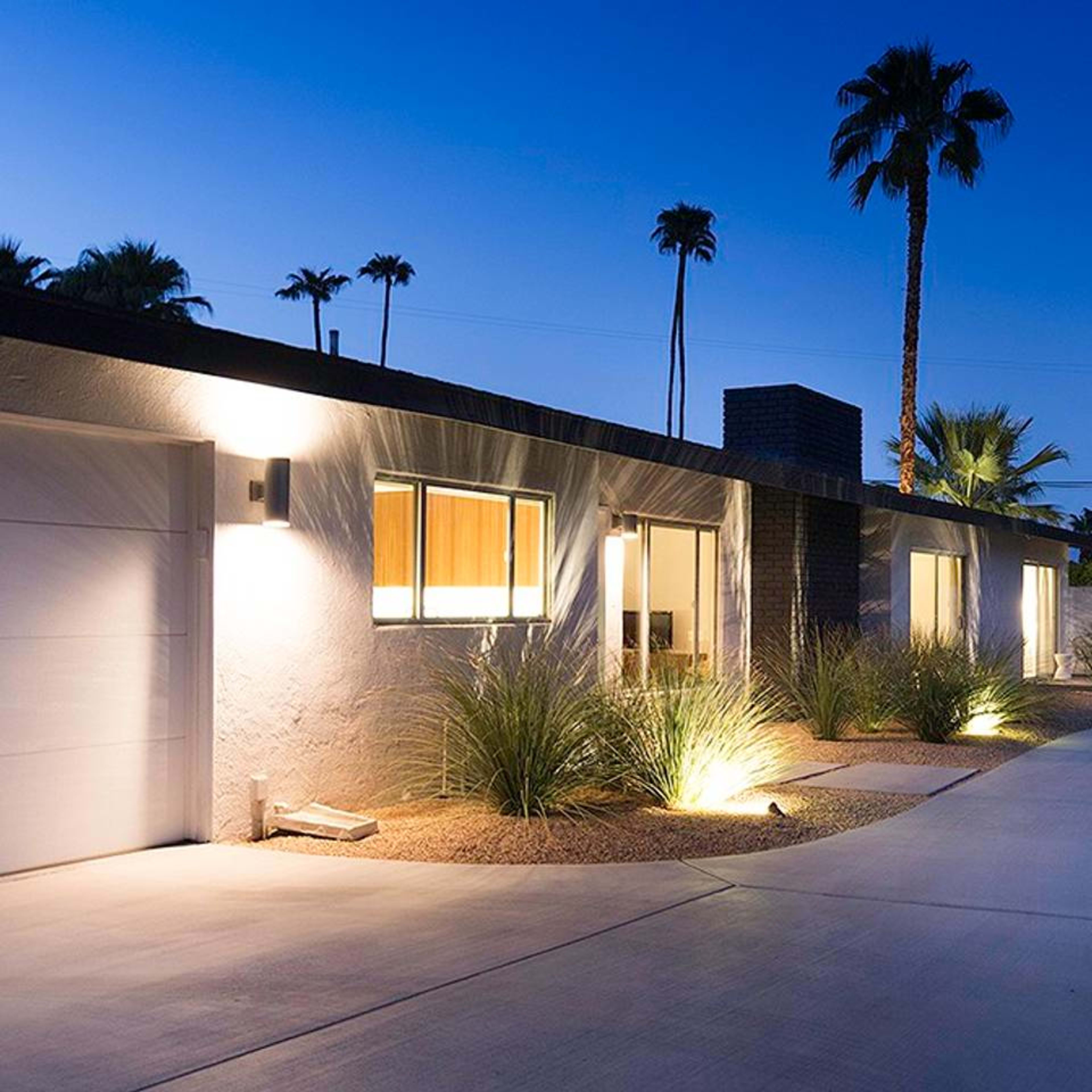A modern single-story house with a landscaped driveway is illuminated by outdoor lighting, surrounded by palm trees at dusk.
