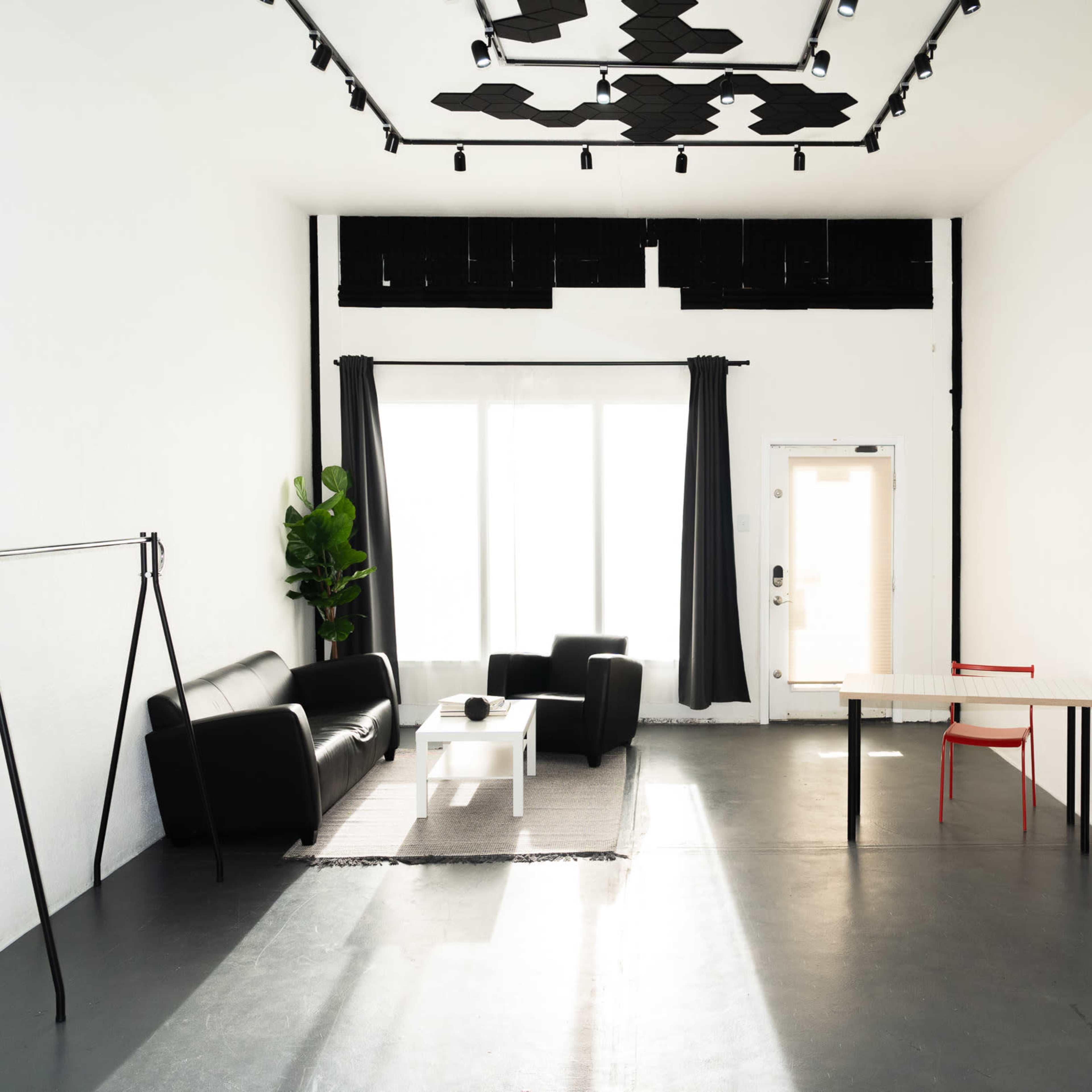A minimalist room with a black sofa, a white coffee table, a large mirror, and a table with a red chair, illuminated by natural light from a large window.