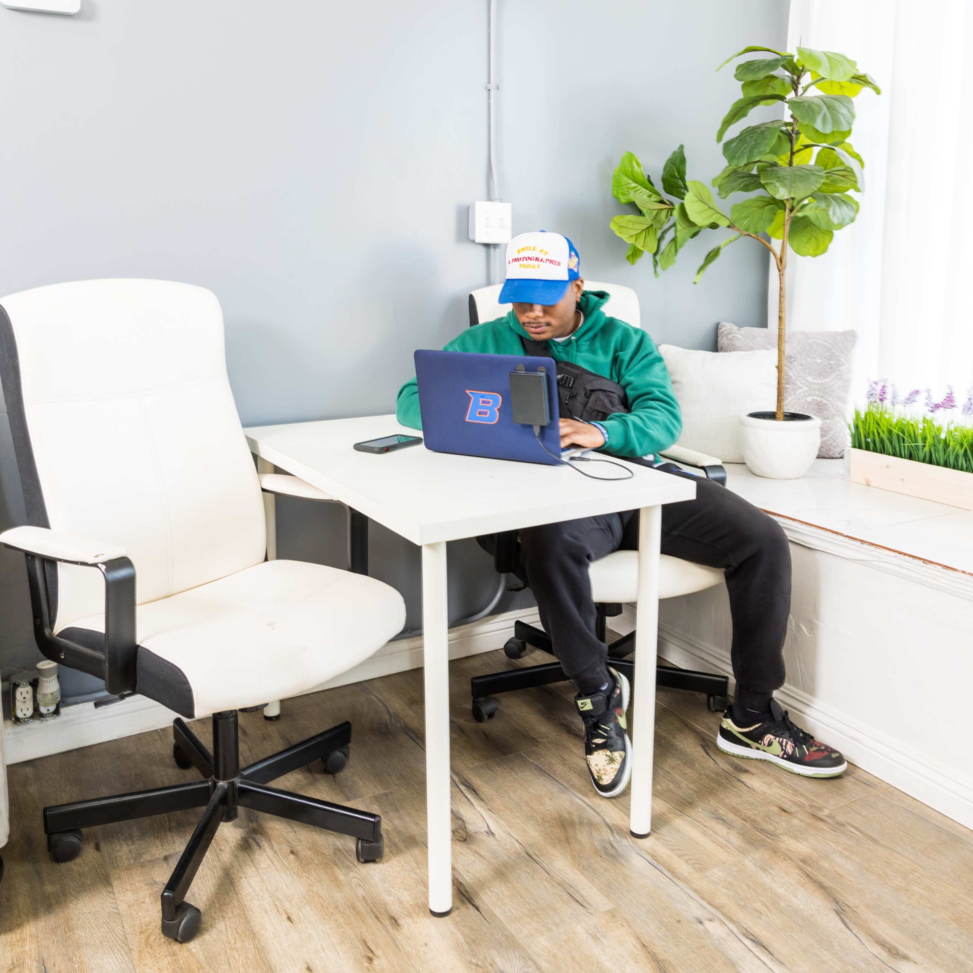 A person wearing a cap sits at a desk with a laptop in a bright office space featuring a potted plant.