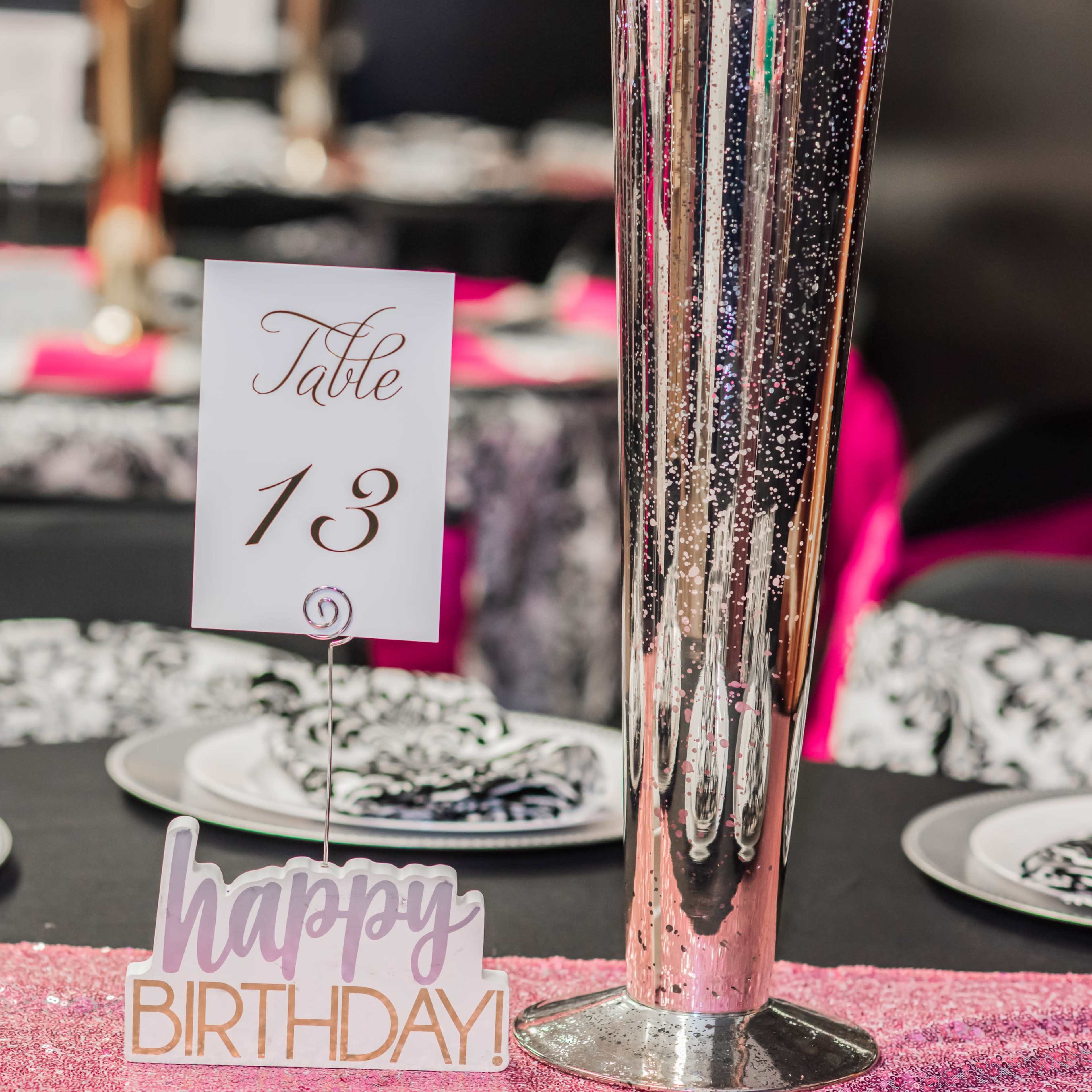 A decorated table setting features a centerpiece, a card displaying "Table 13," and a sign that reads "happy BIRTHDAY."