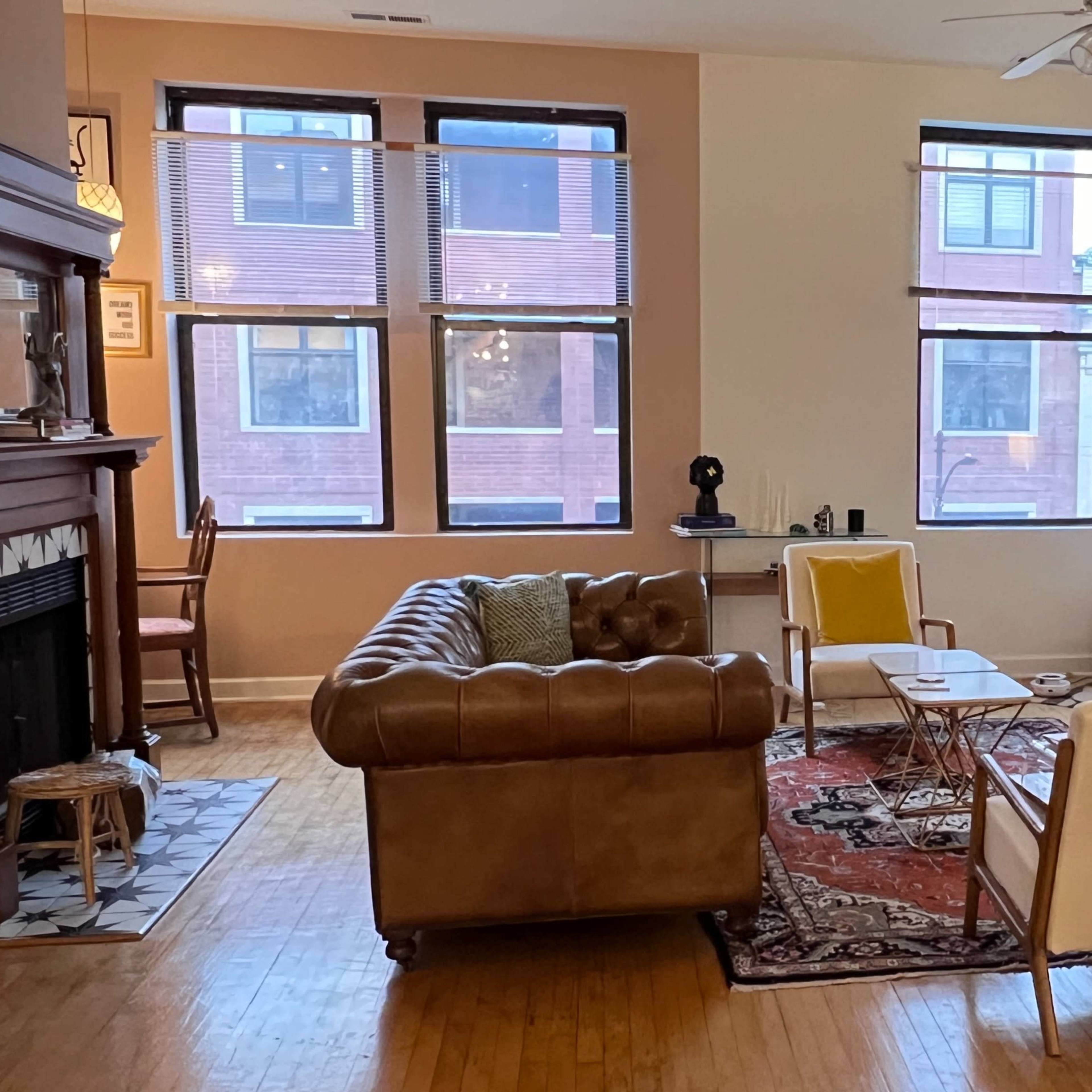A spacious living room with large windows, a brown leather couch, a white media console with a television, and a mix of seating arrangements.