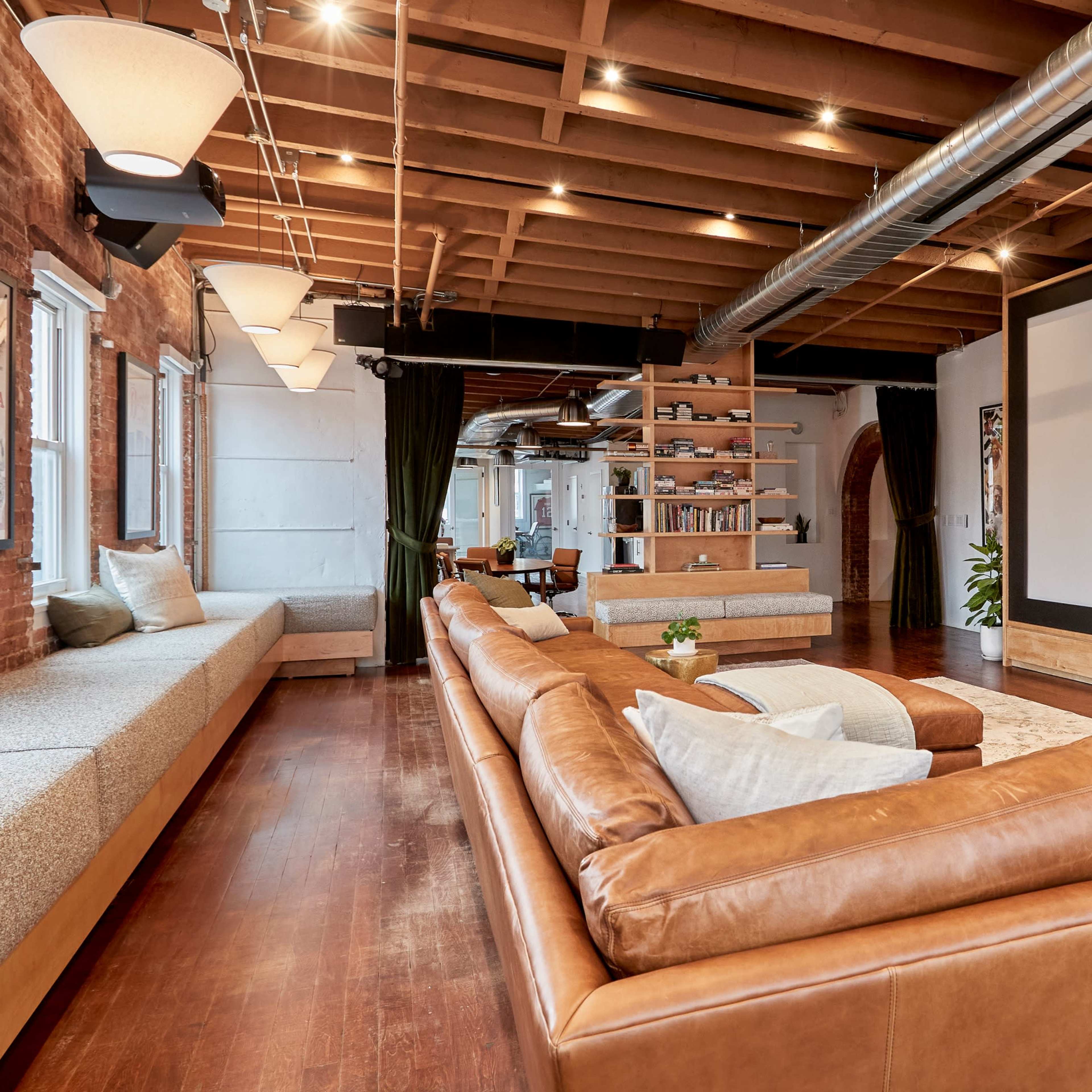 A spacious living room featuring a large brown leather sectional sofa, a wall-mounted screen, and a cozy seating area beneath windows with wooden beams overhead.