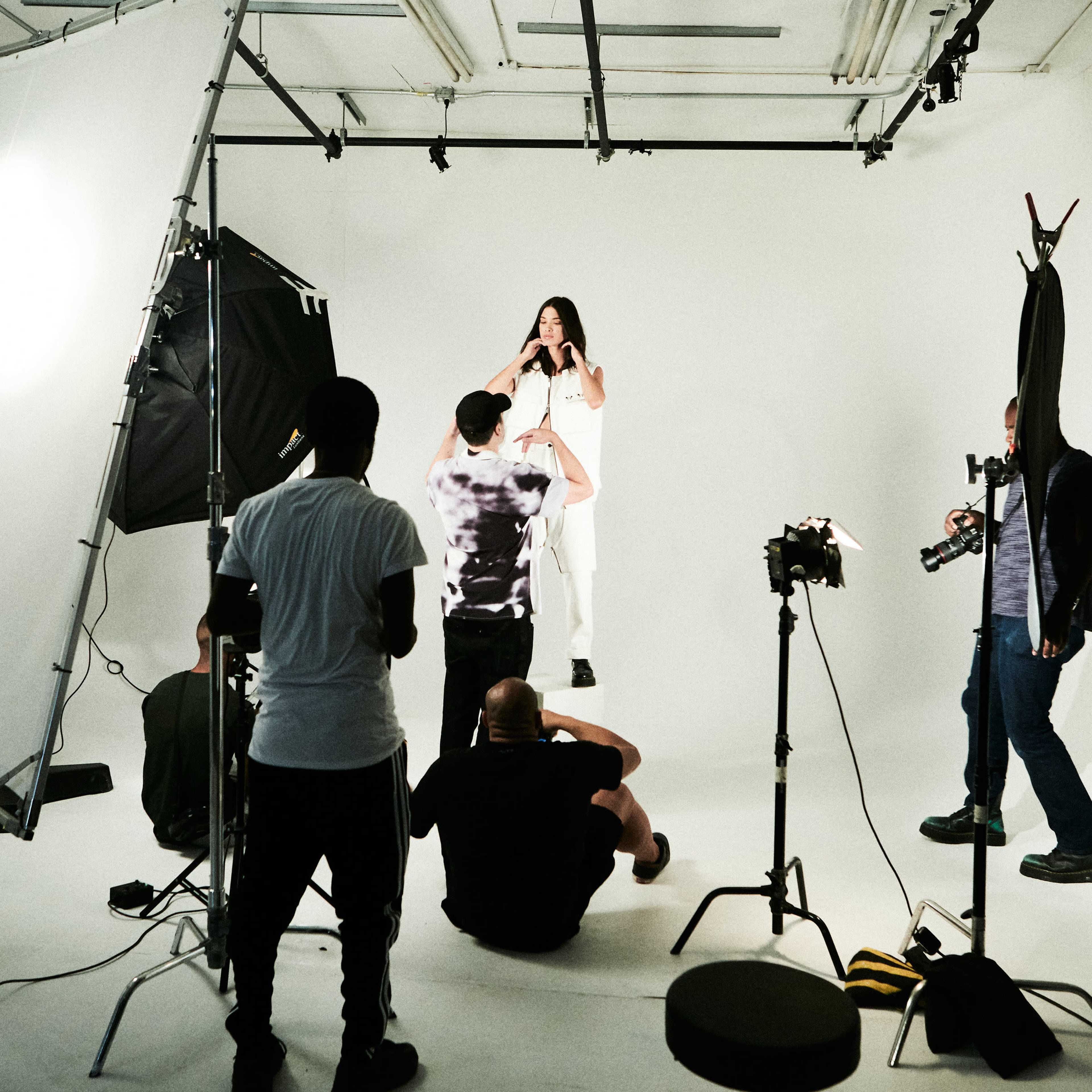A group of photographers and crew members are working in a studio with a model posing against a white backdrop.