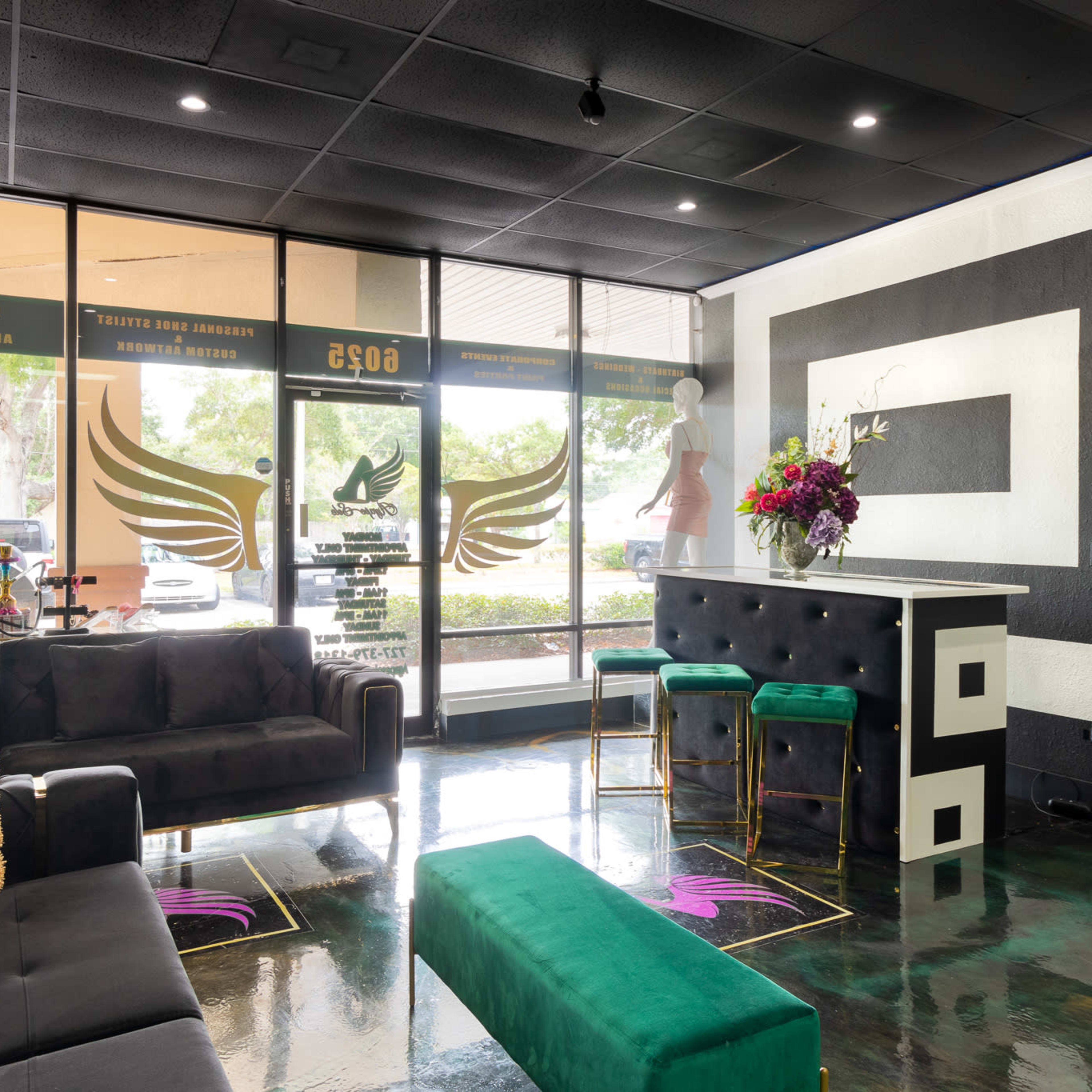 The interior of a modern salon features black sofas, a green bench, a stylish reception area with high stools, and large windows showcasing outdoor views.
