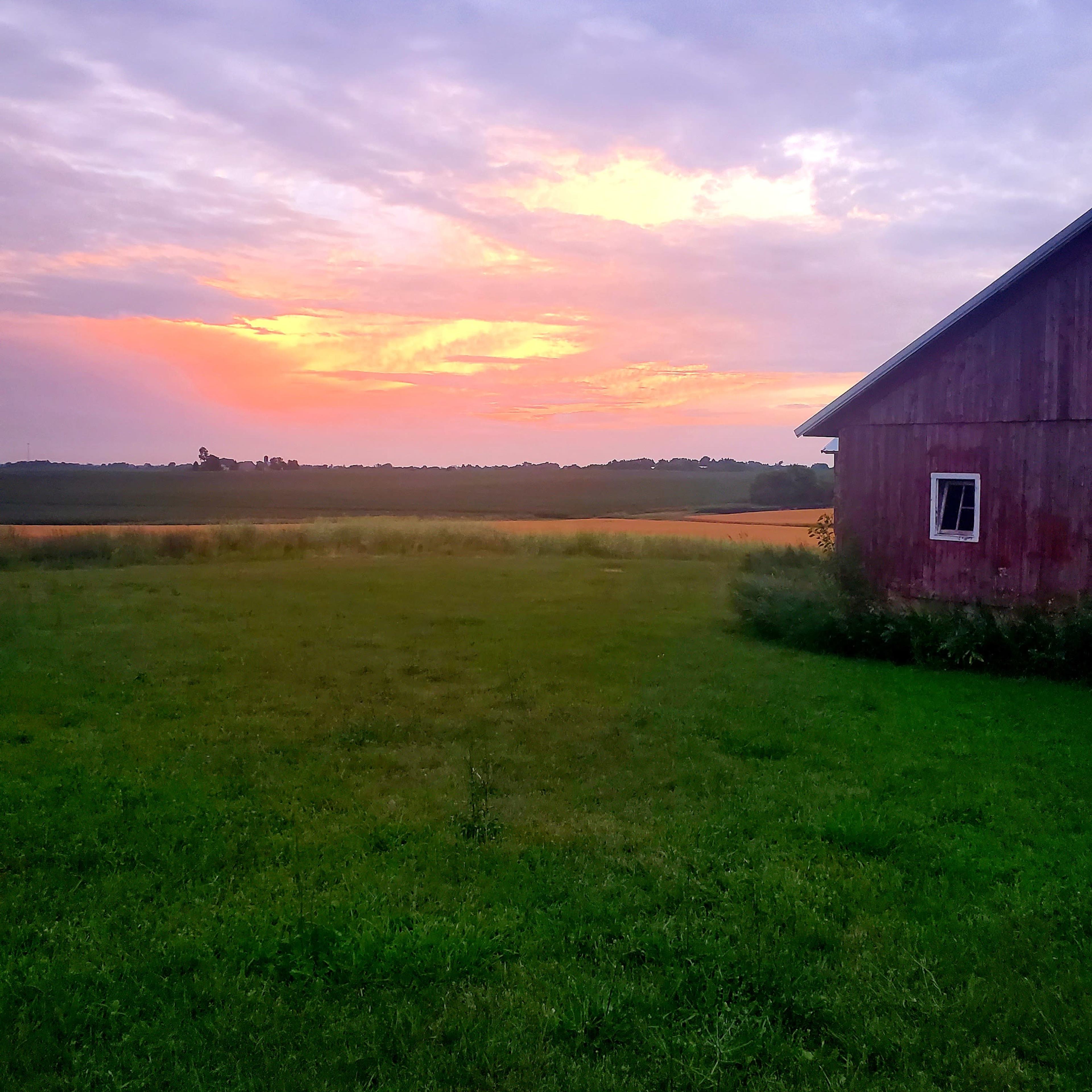 A red barn stands on green grass under a colorful sky at sunset, with fields stretching into the distance.