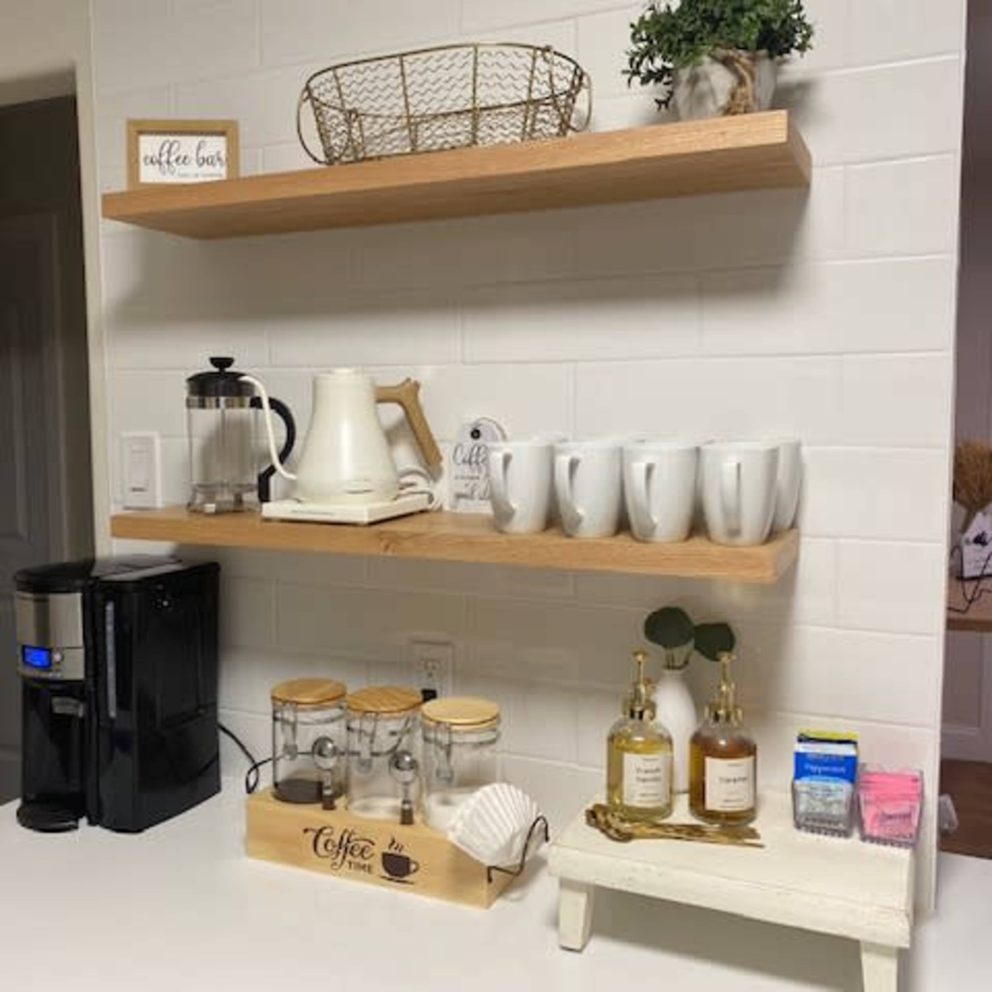 A coffee station features a coffee maker, several mugs, a French press, and a decorative tray with coffee supplies on a countertop beneath wooden shelves.