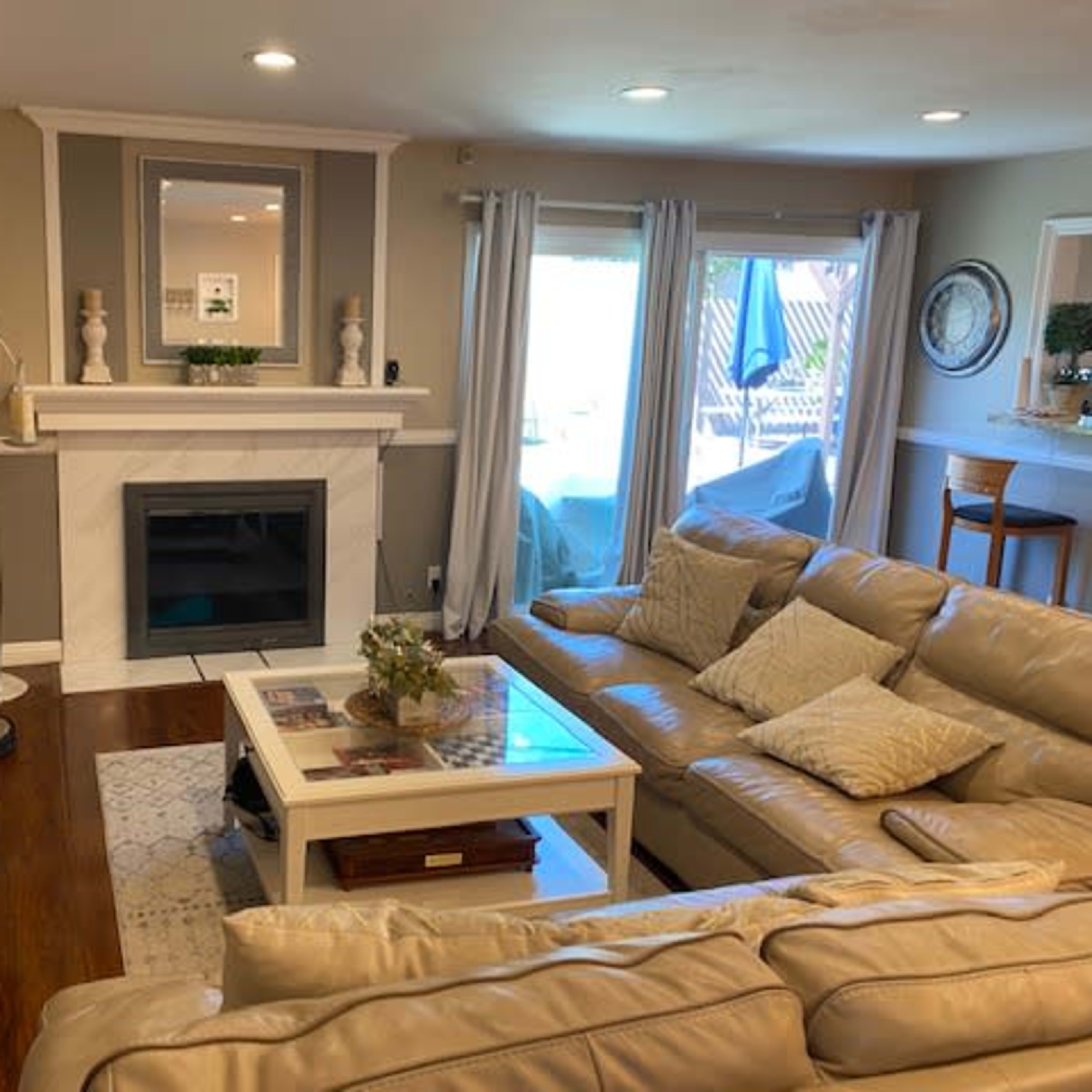 A living room with a cream-colored leather sectional sofa, a coffee table, a wall-mounted TV, and a white marble fireplace, alongside large windows leading to an outdoor patio.