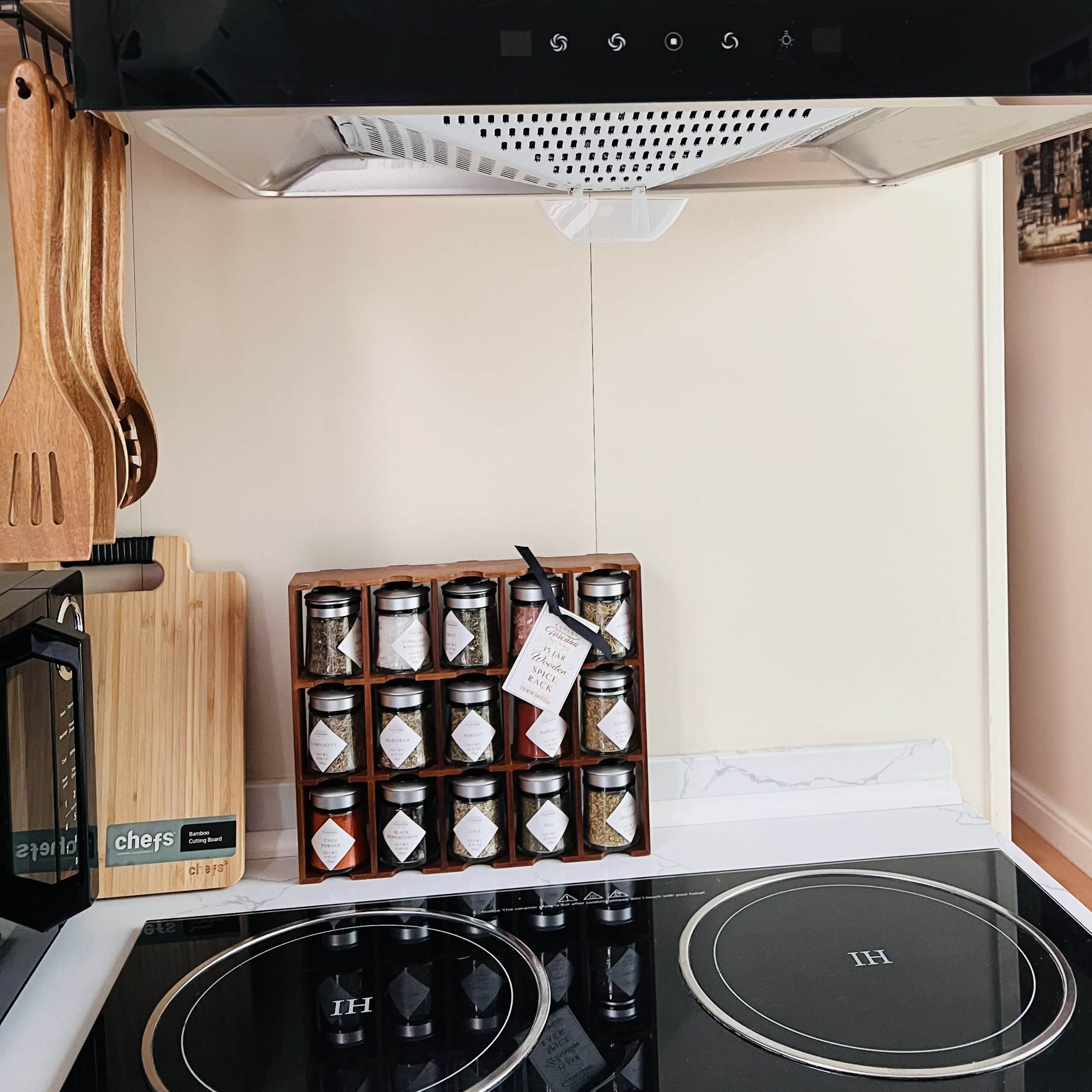 A kitchen countertop with an induction cooktop, a wooden cutting board, wooden utensils, and a rack of spice jars labeled with tags.