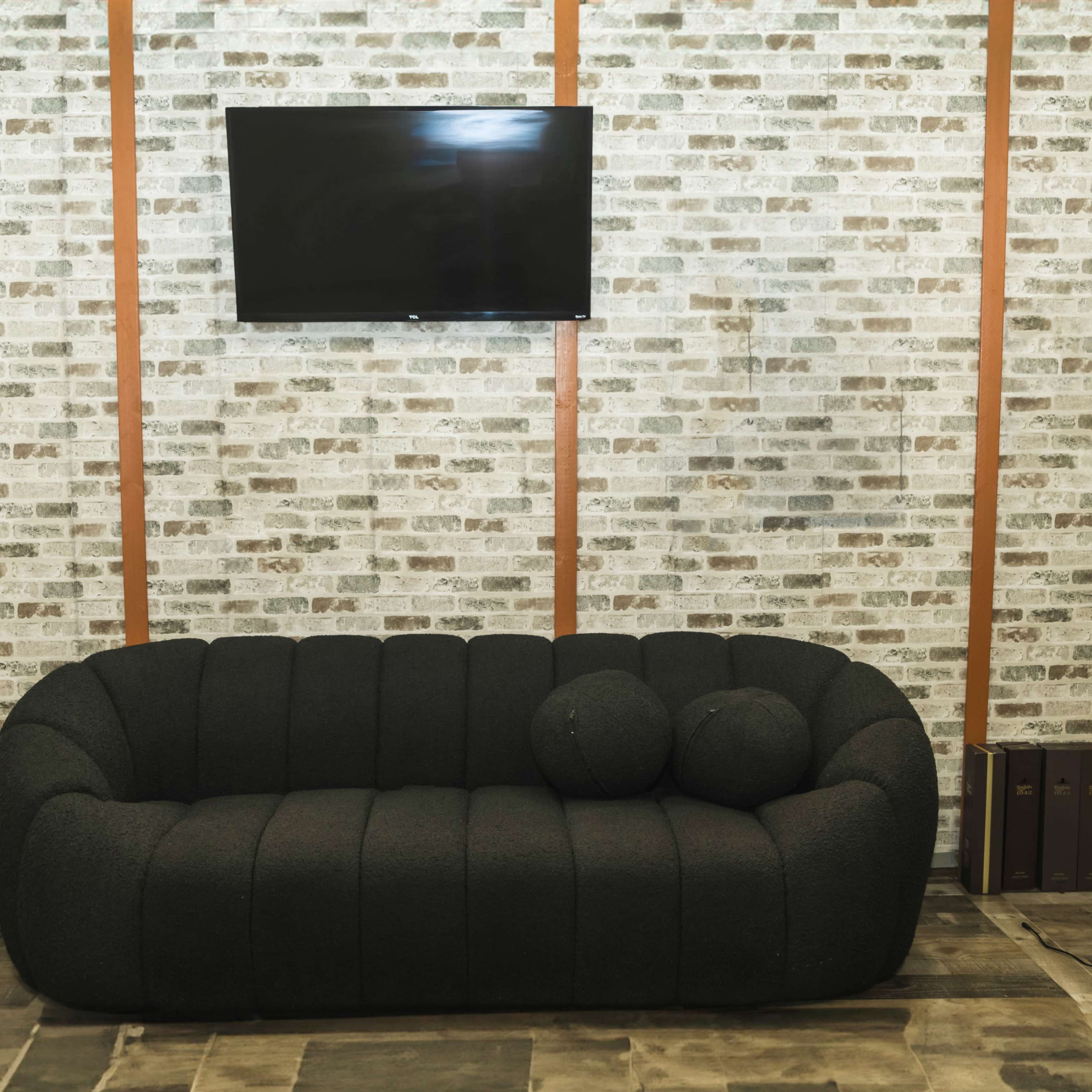 A black upholstered sofa with two matching pillows is positioned in front of a flat-screen television mounted on a brick-patterned wall.