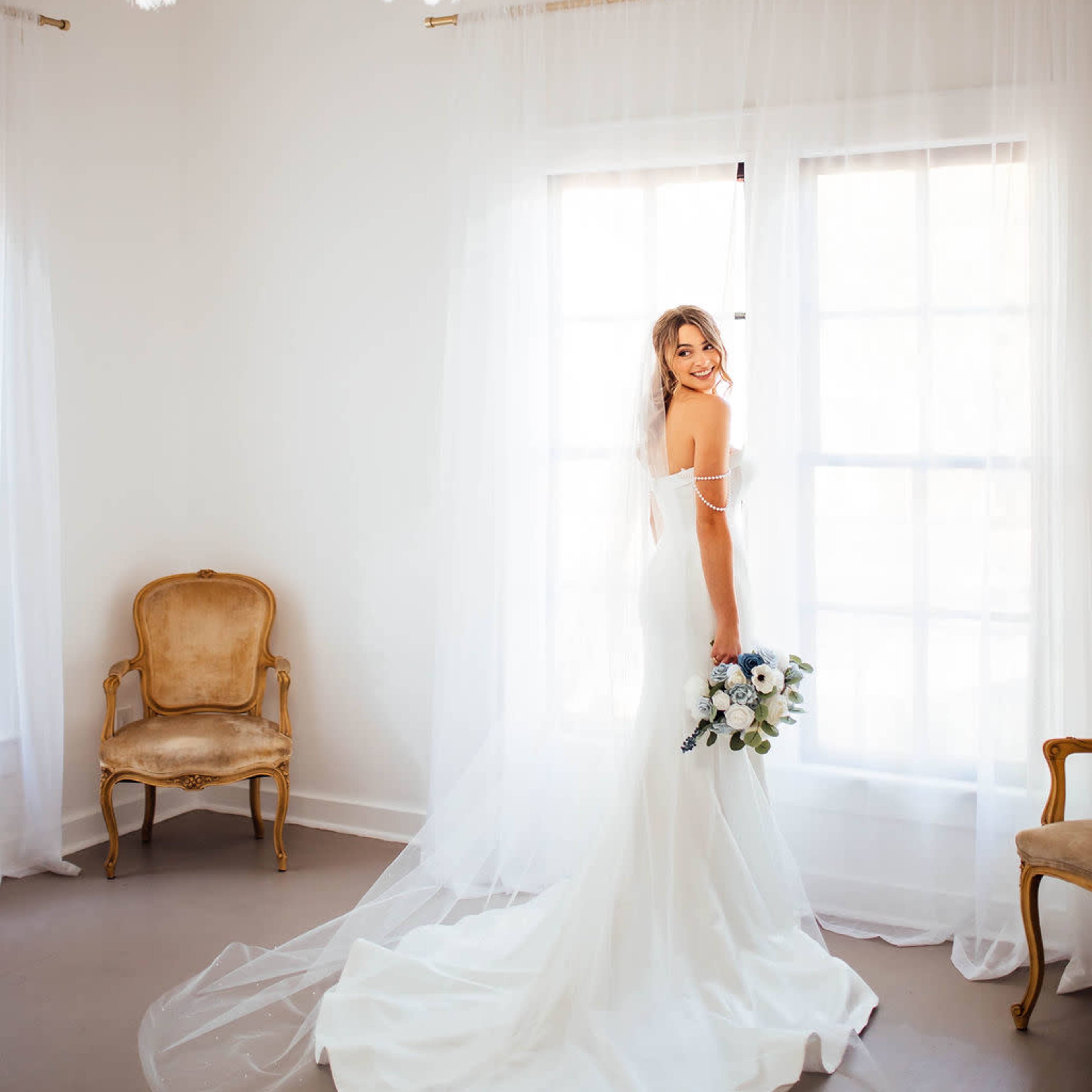 A bride stands by a large window, holding a bouquet, with sheer curtains draping around her in a softly lit room.