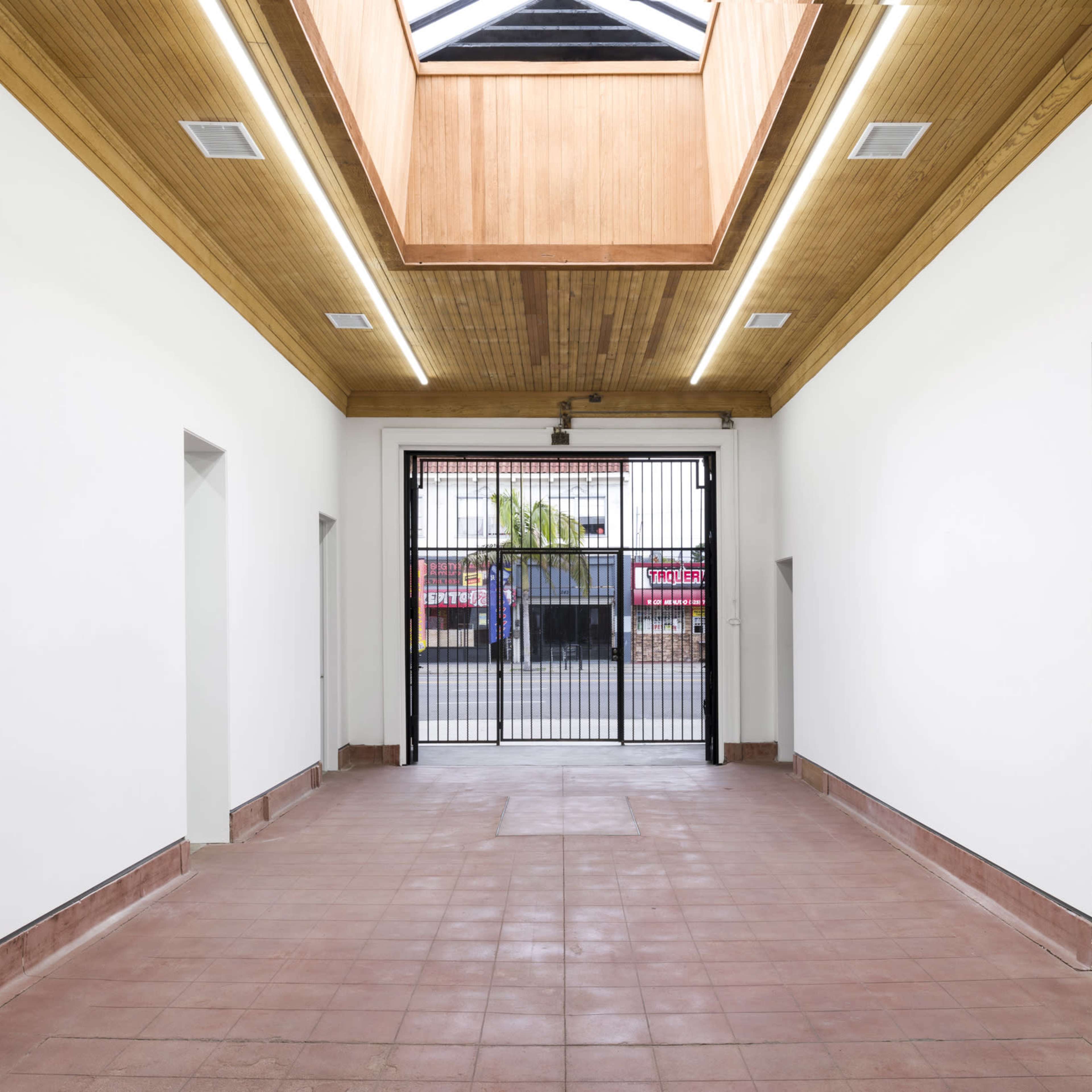 A spacious, empty interior with a wooden ceiling and a large skylight, leading to an open gate facing a street.