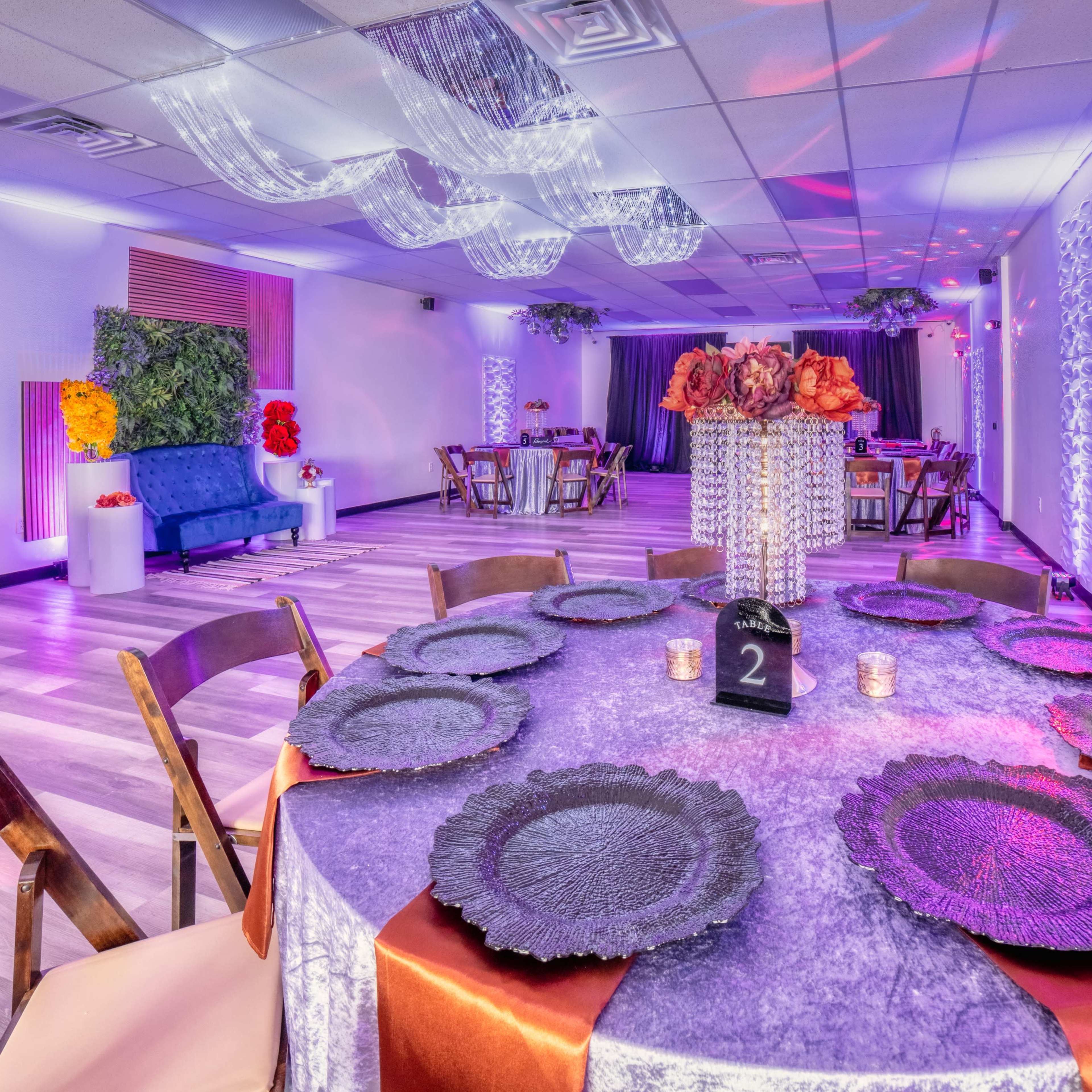 A beautifully arranged event space features round tables with decorative tableware, a blue couch, and floral centerpieces under colorful lighting.
