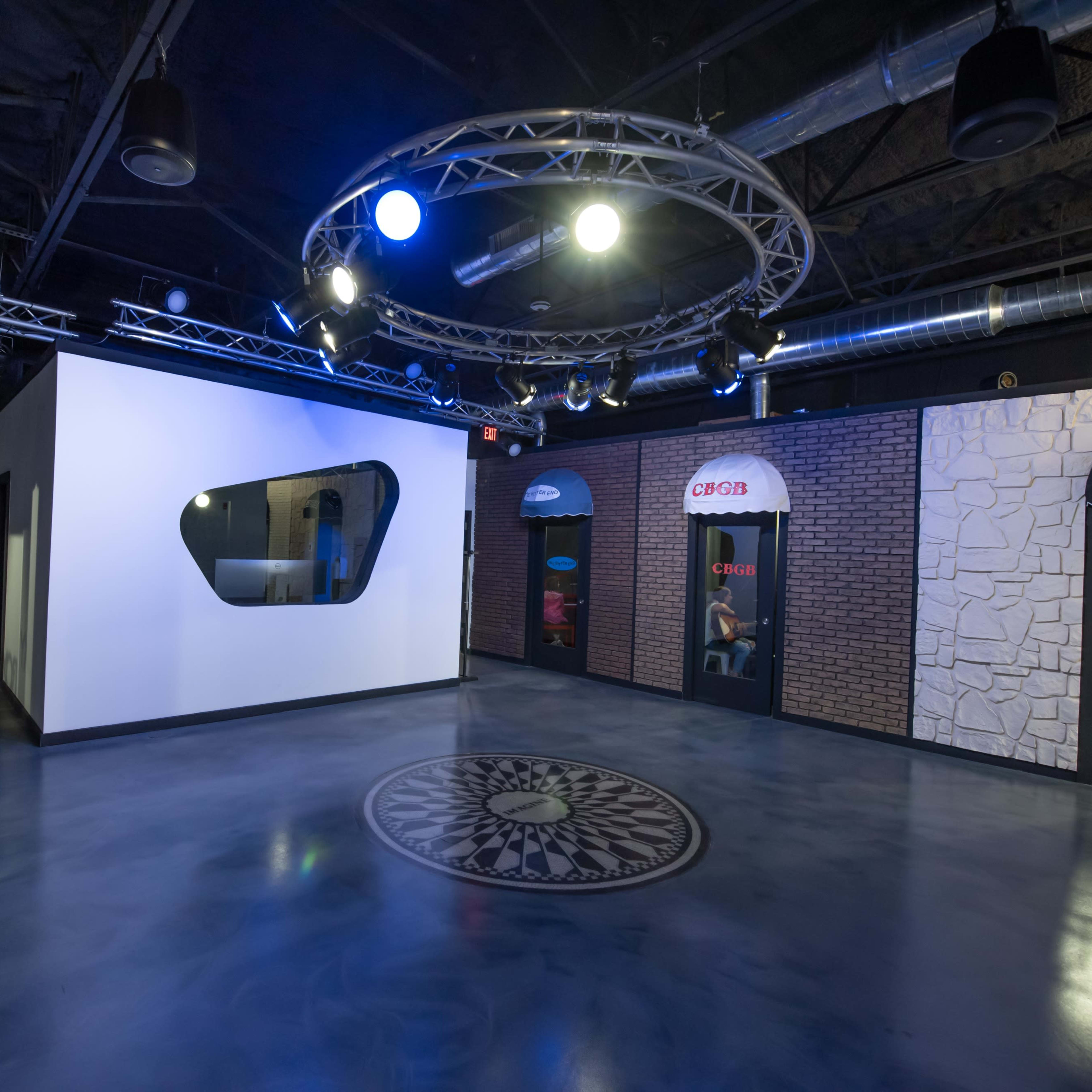 A modern indoor space with a circular design feature on the floor, illuminated by overhead lights, and flanked by doorways resembling storefronts.