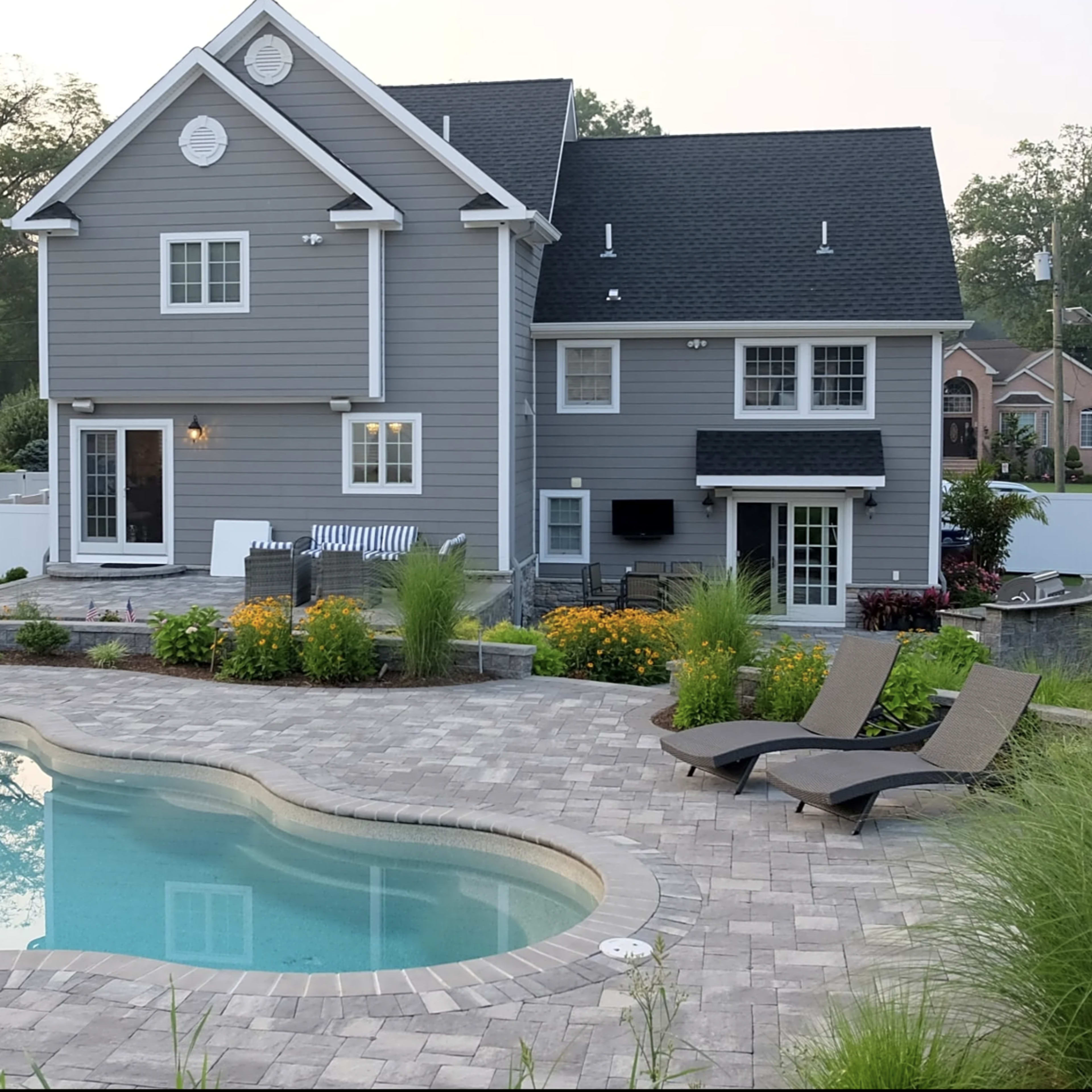 A spacious gray house with a landscaped yard features a swimming pool surrounded by lounge chairs and flower beds.