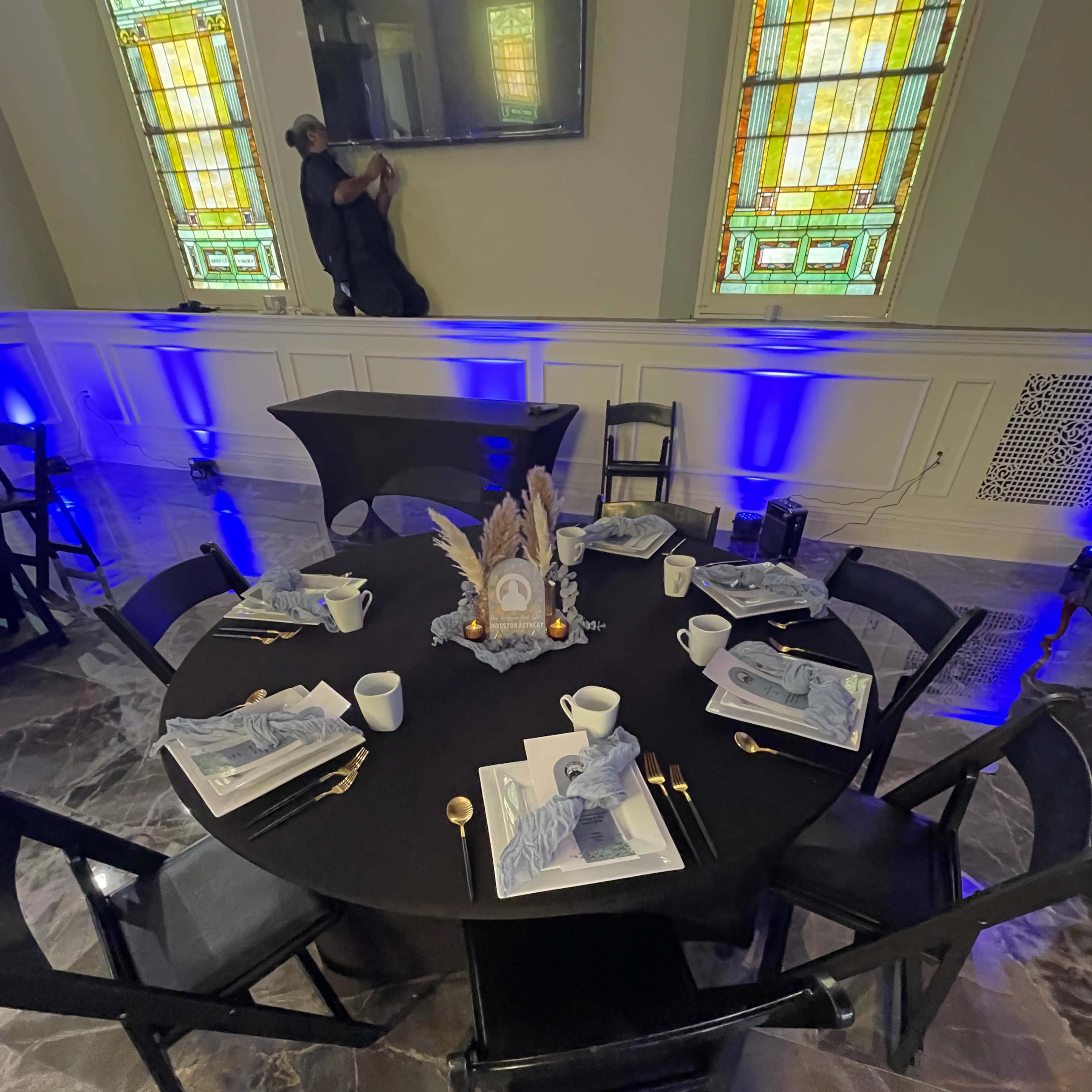A round black table is elegantly set with gray-themed dinnerware, gold utensils, and decorative accents, surrounded by black chairs in a well-lit room with stained glass windows.