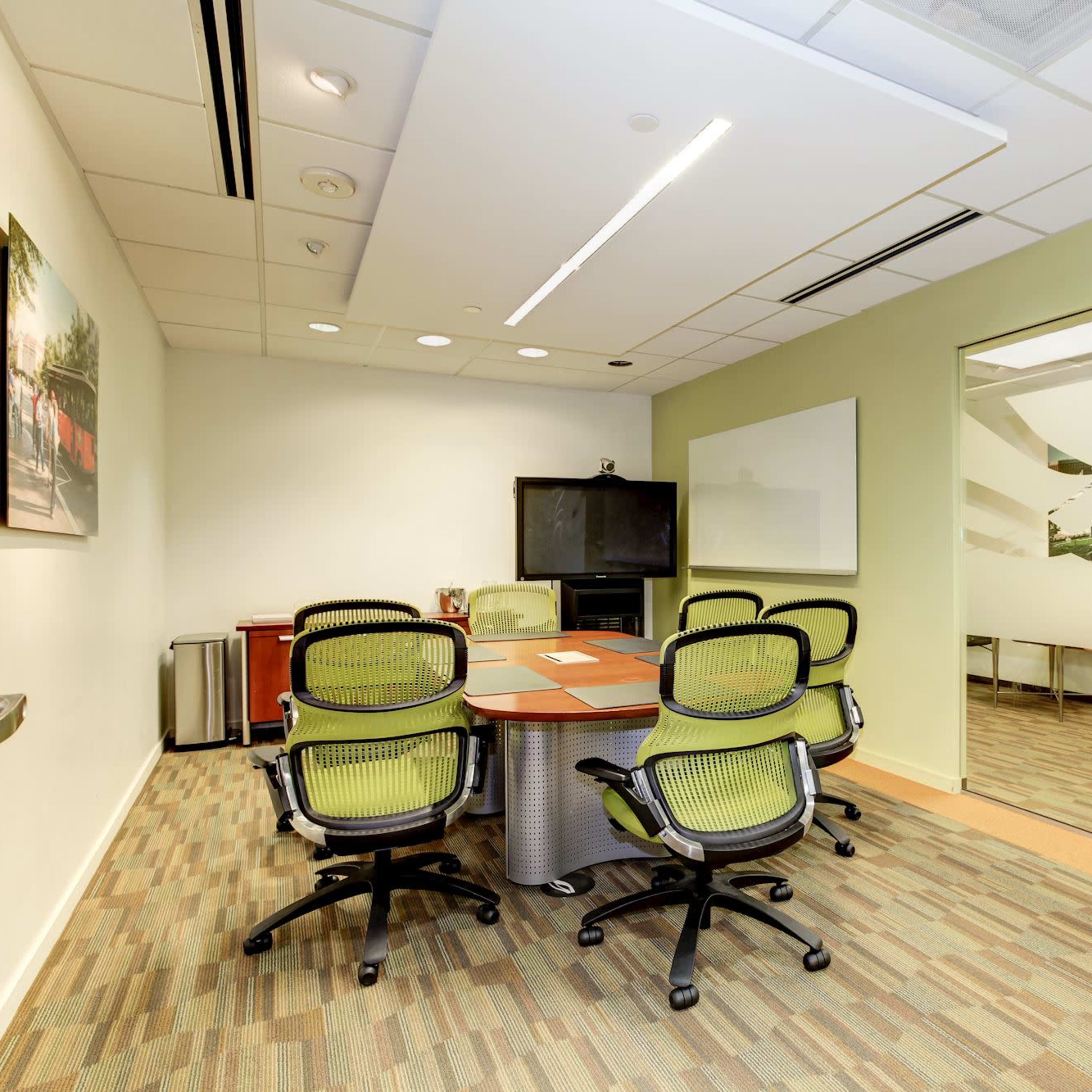 A modern conference room with a central table surrounded by green ergonomic chairs, a glass wall, and a television on one side.