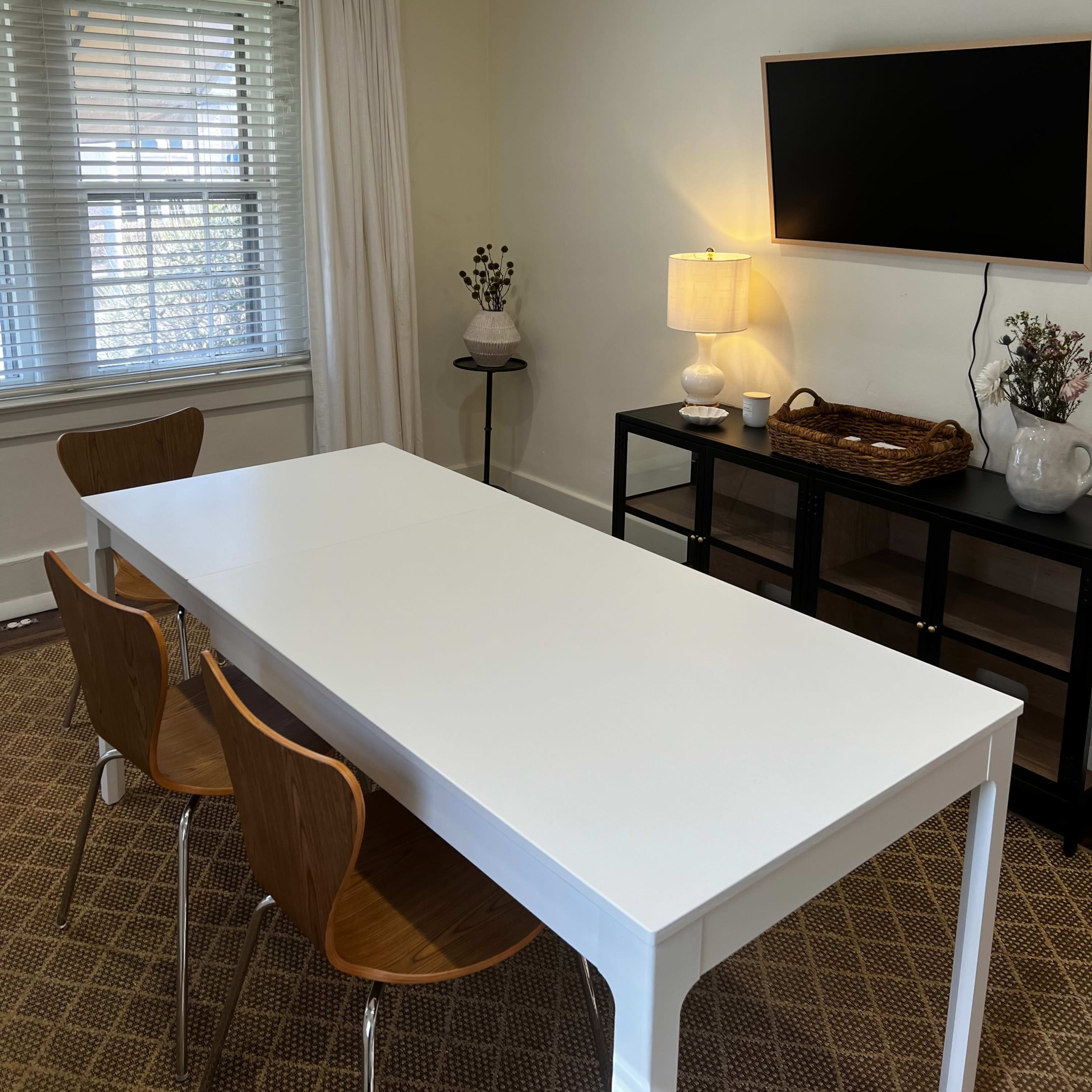 A minimalist dining area features a white table with three wooden chairs, a television on the wall, and a lamp on a side table.