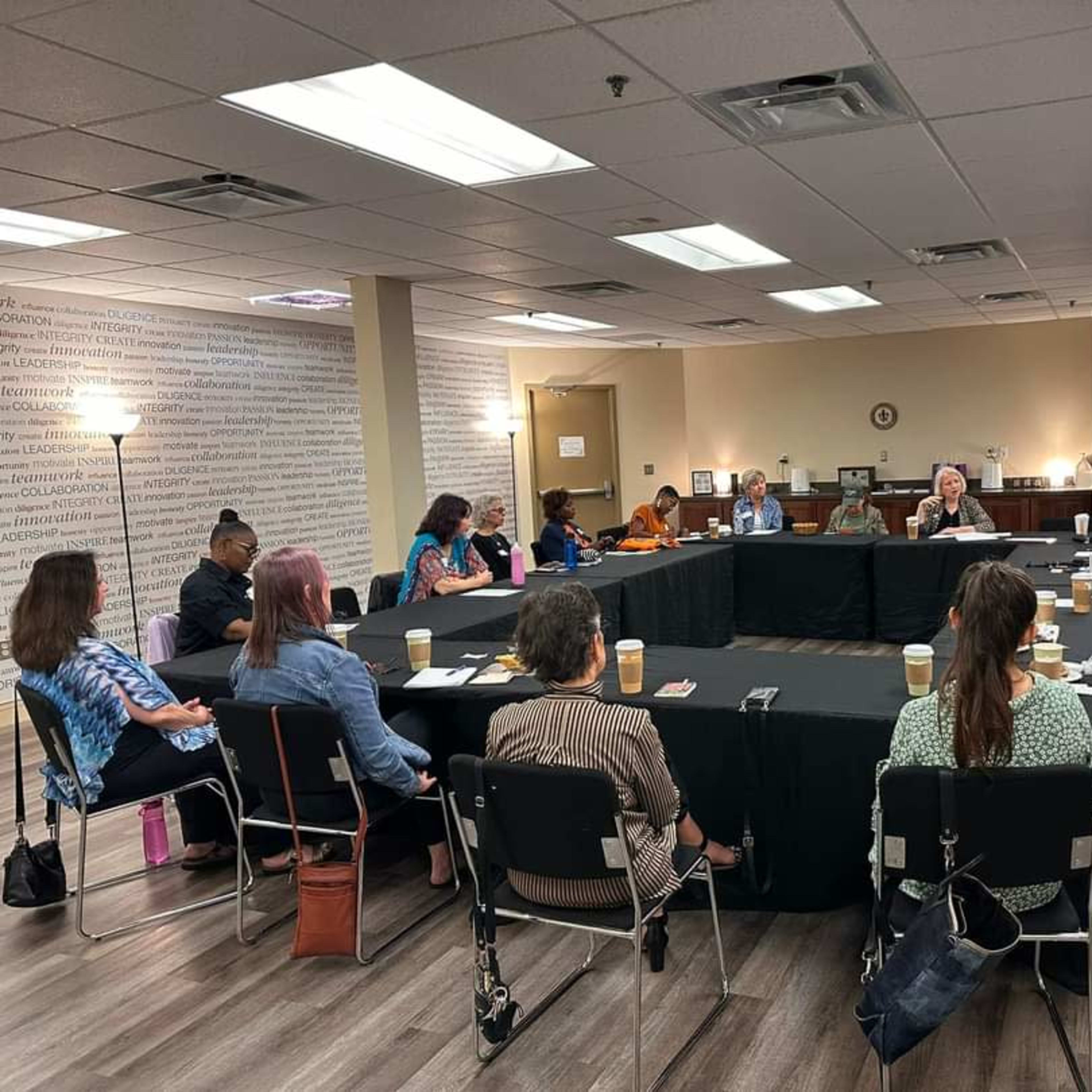 A group of people sits around a large table in a meeting room, engaged in discussion.
