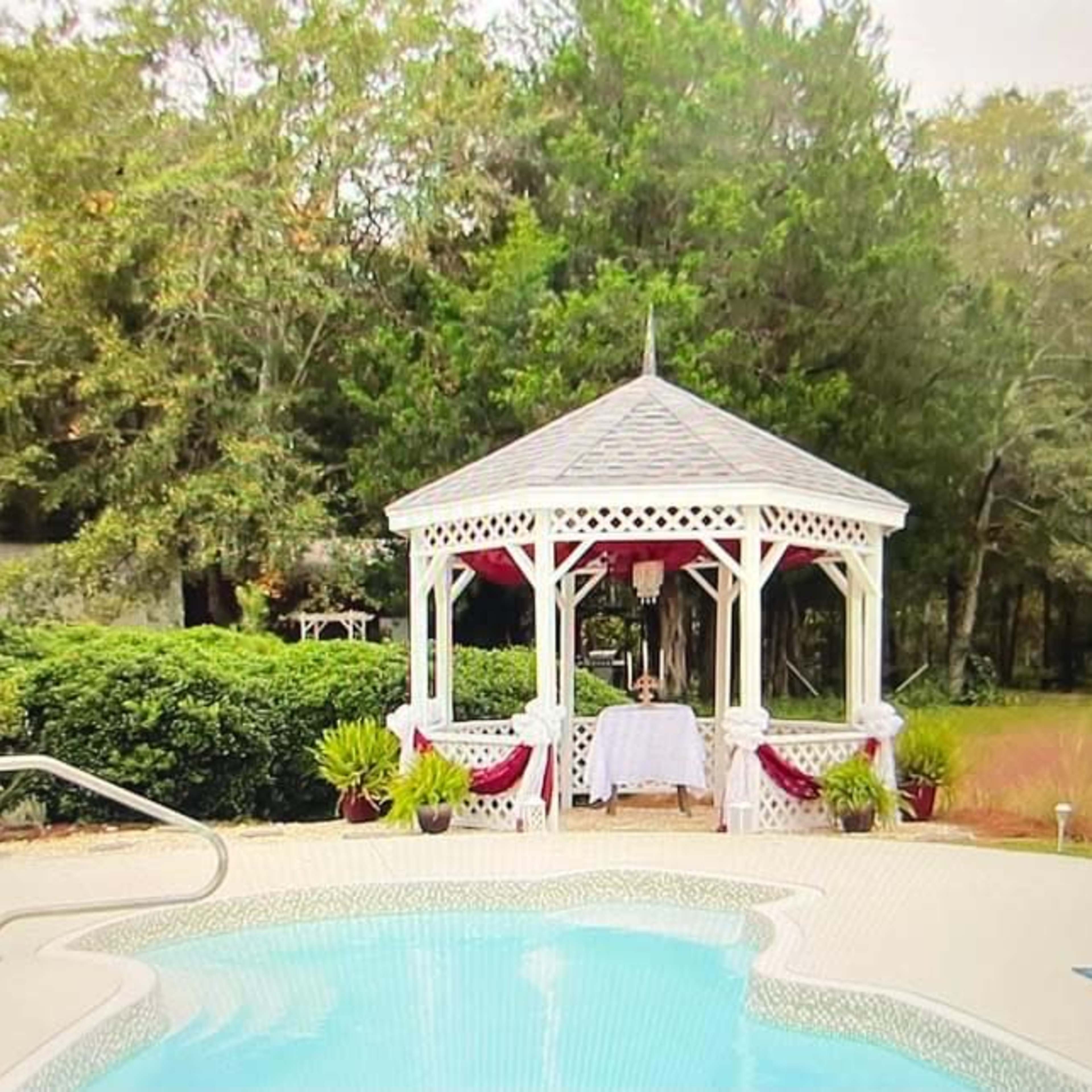A white gazebo with red accents is situated near a sparkling pool, surrounded by greenery.