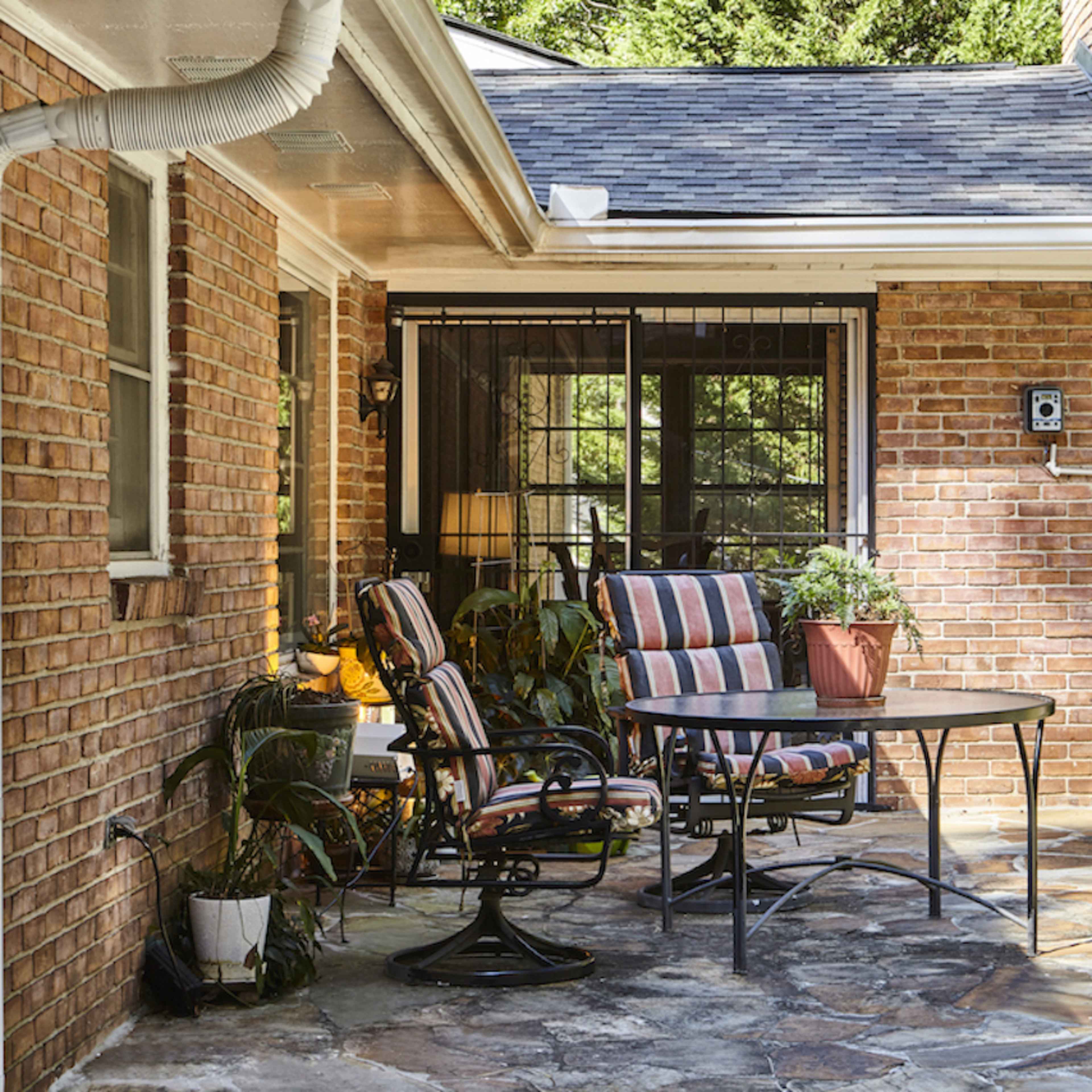 A patio area features a round table with striped chairs, surrounded by greenery and blooming flowers, attached to a brick house.