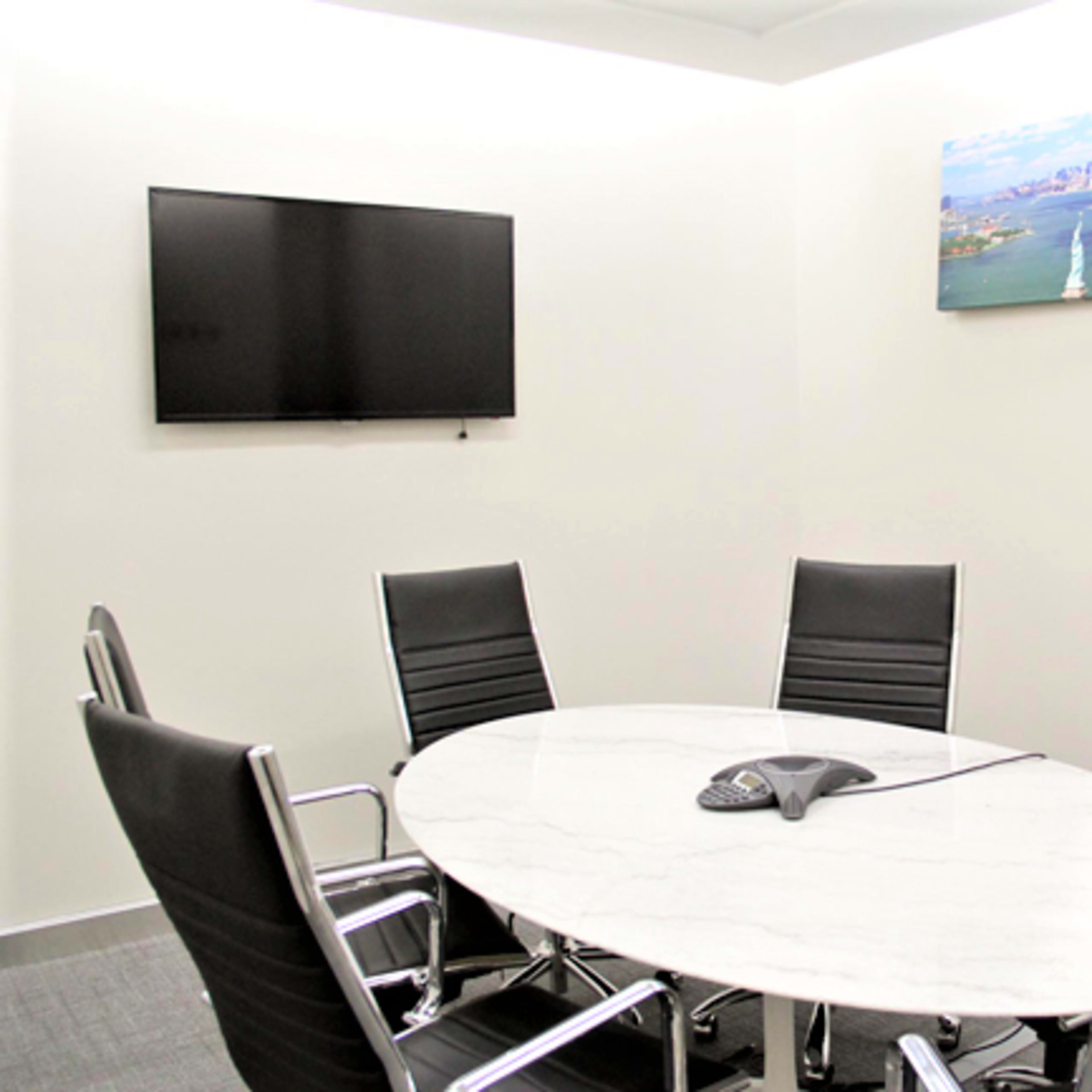 A modern conference room with a round marble table, six black chairs, a wall-mounted TV, and a landscape painting of a city view.