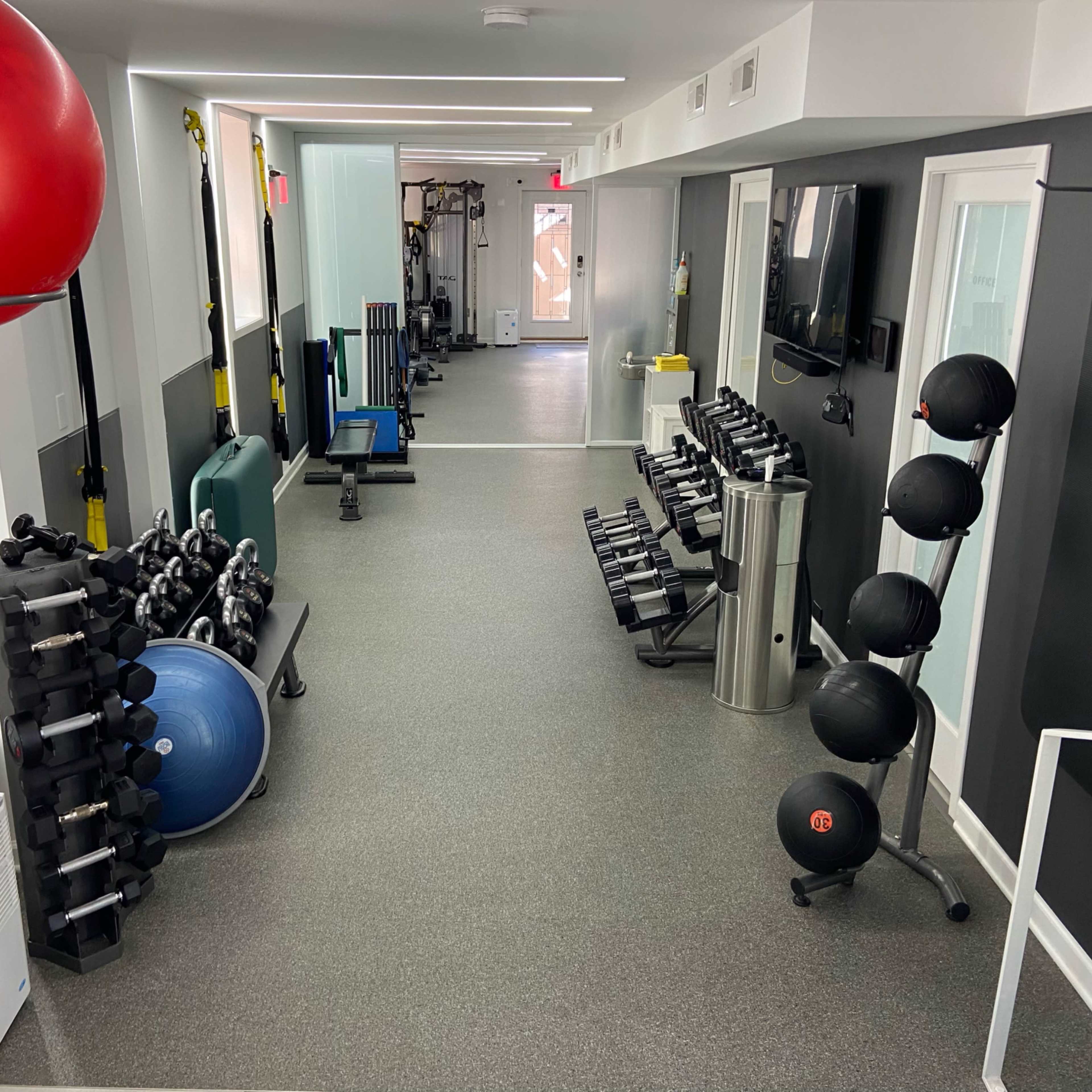 A fitness gym with various equipment, including dumbbells, a stability ball, and a rowing machine, arranged along the walls of a well-lit room.