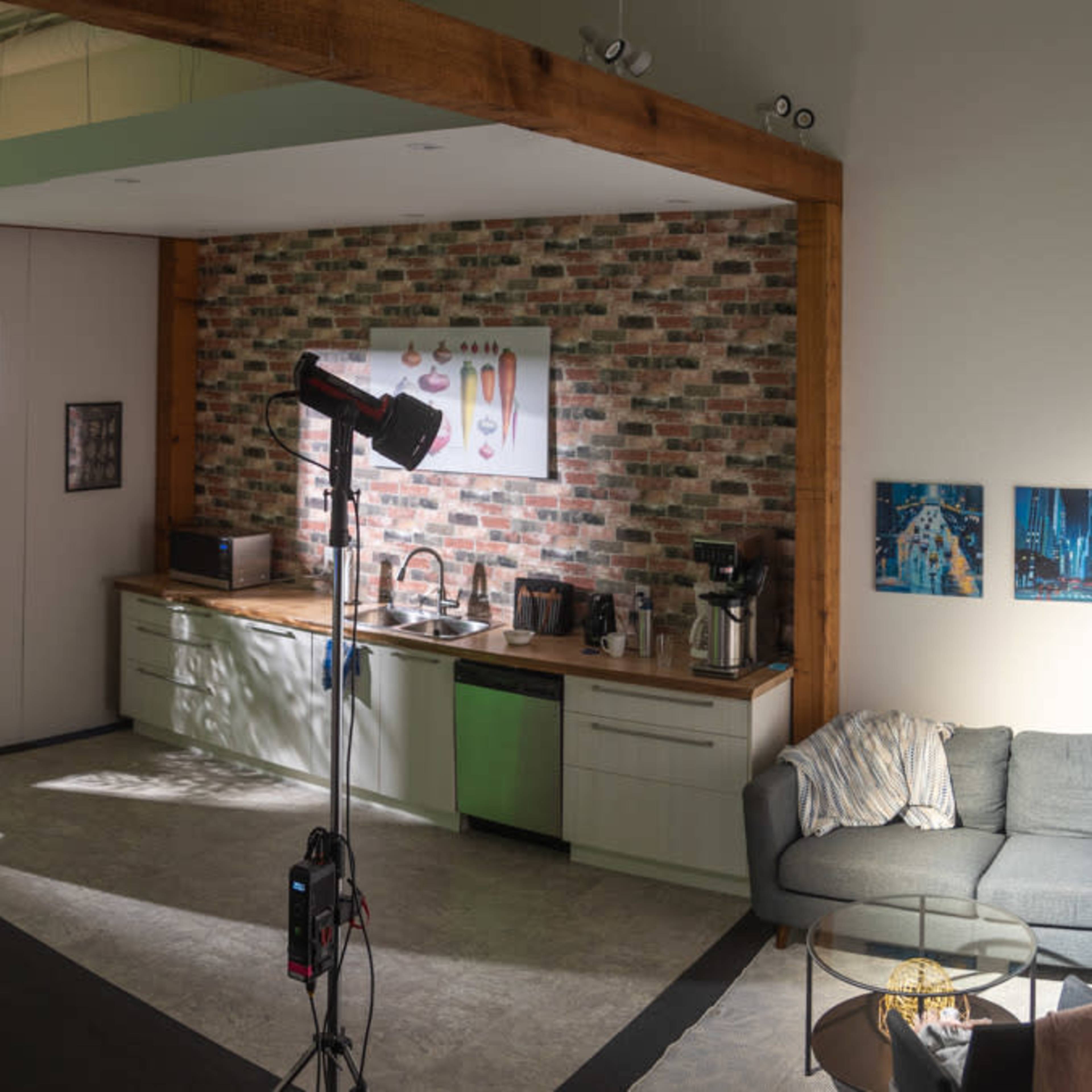 A spacious interior featuring a kitchen area with a brick accent wall, a seating area with a couch, and studio lighting equipment set up for a photo or video shoot.