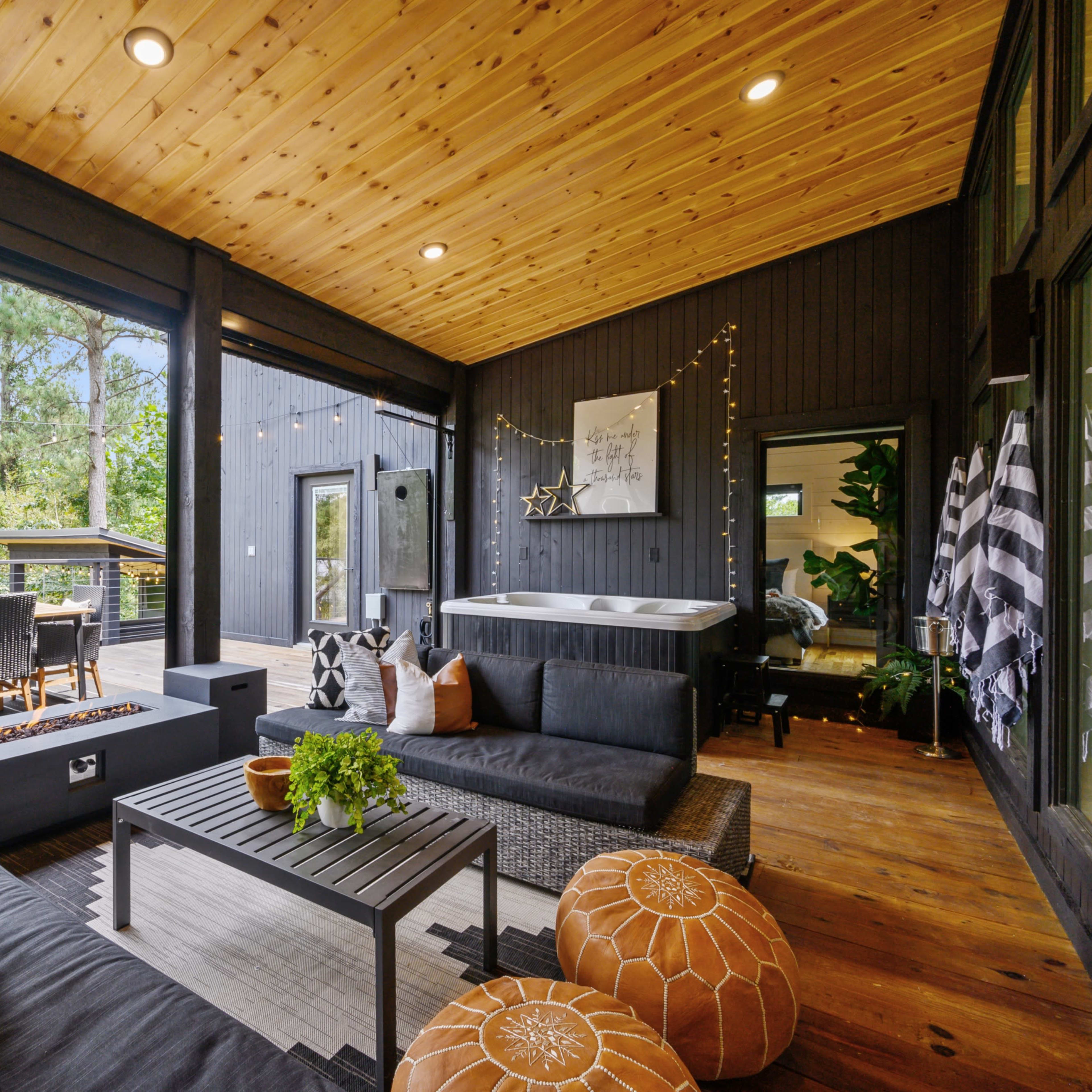 A covered outdoor living space featuring a seating area with dark wicker furniture, a hot tub, and a dining table against a backdrop of greenery.