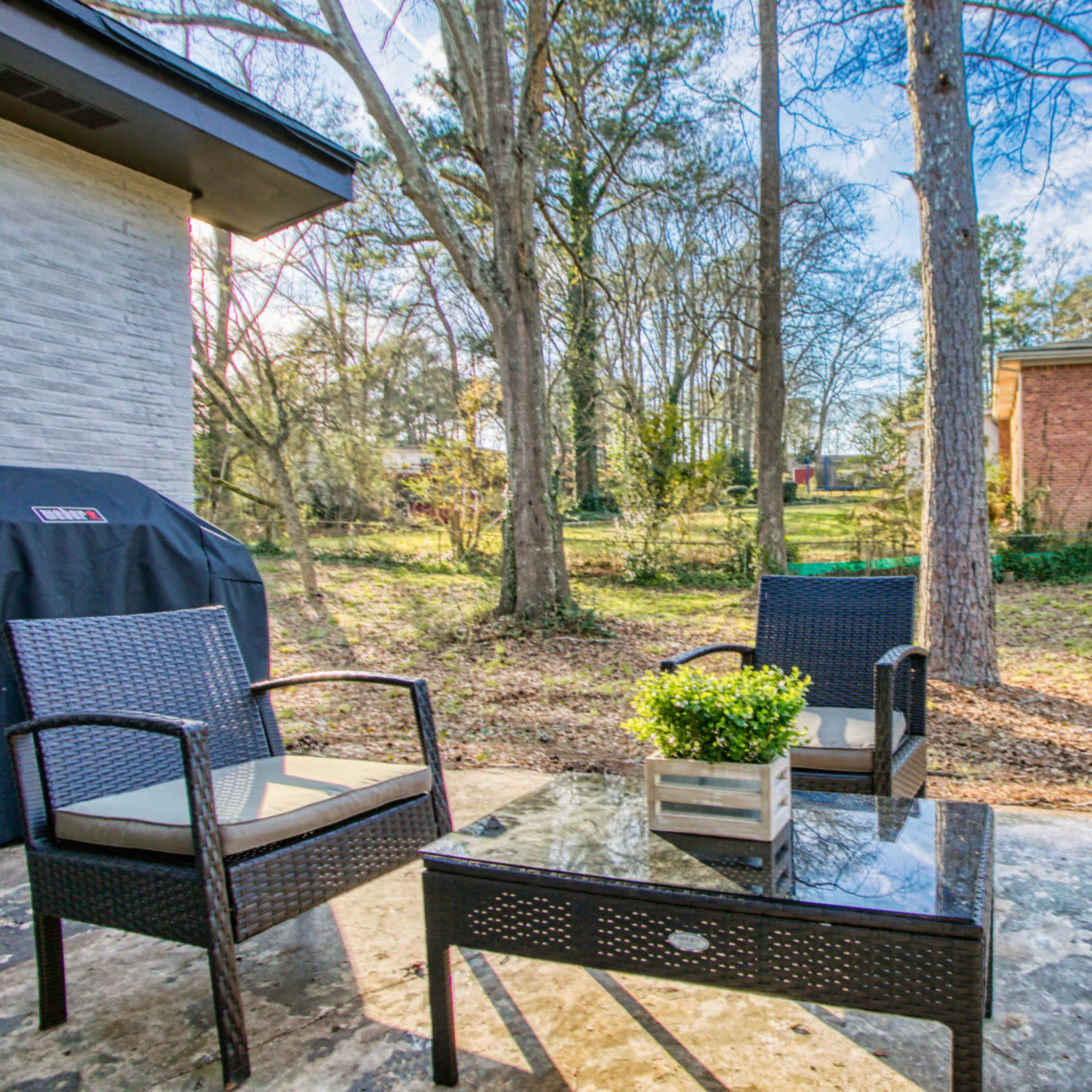A patio area with wicker furniture and a glass table overlooks a grassy backyard surrounded by trees.