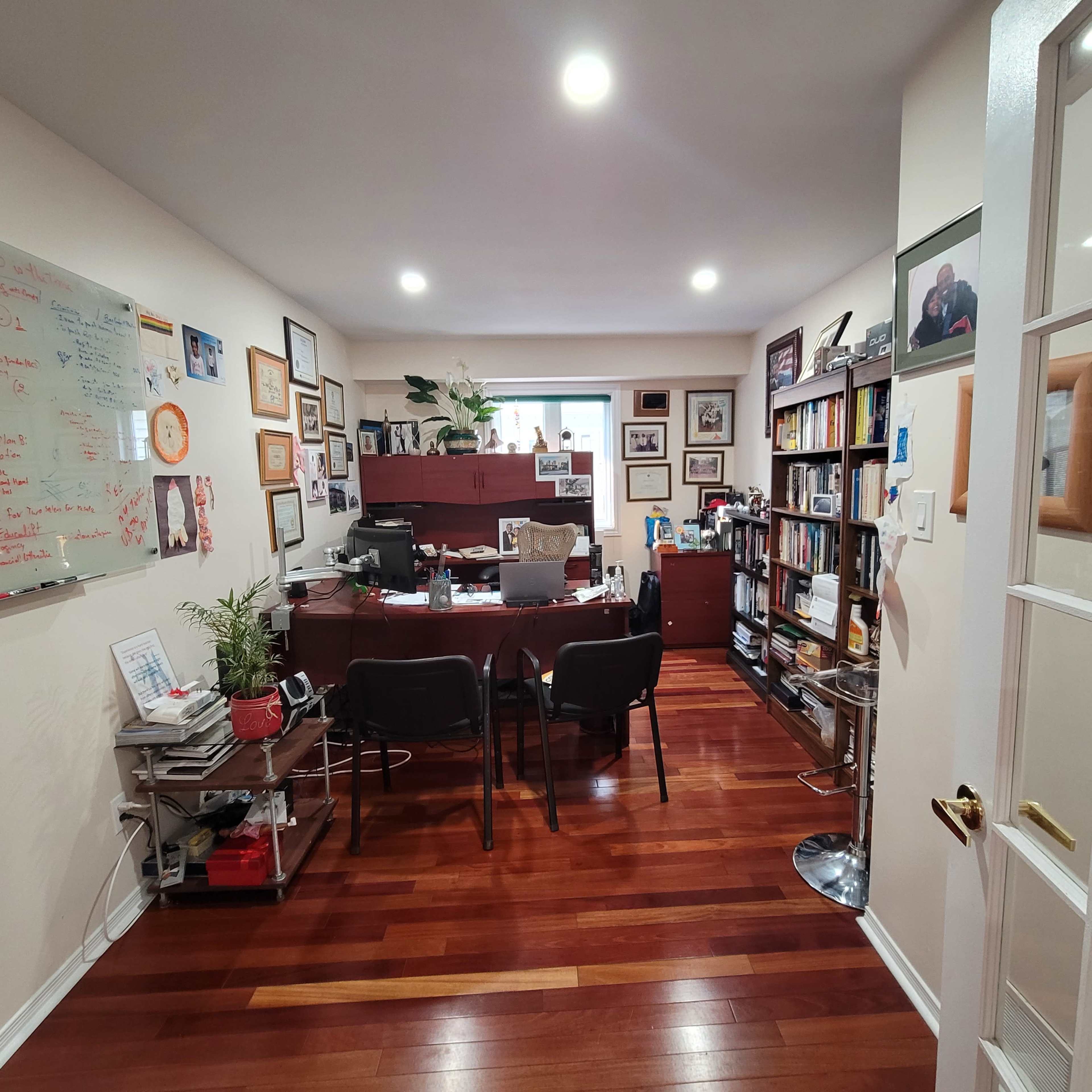 An office with a wooden desk, chairs, shelves lined with books, and walls decorated with various framed photographs and a whiteboard.