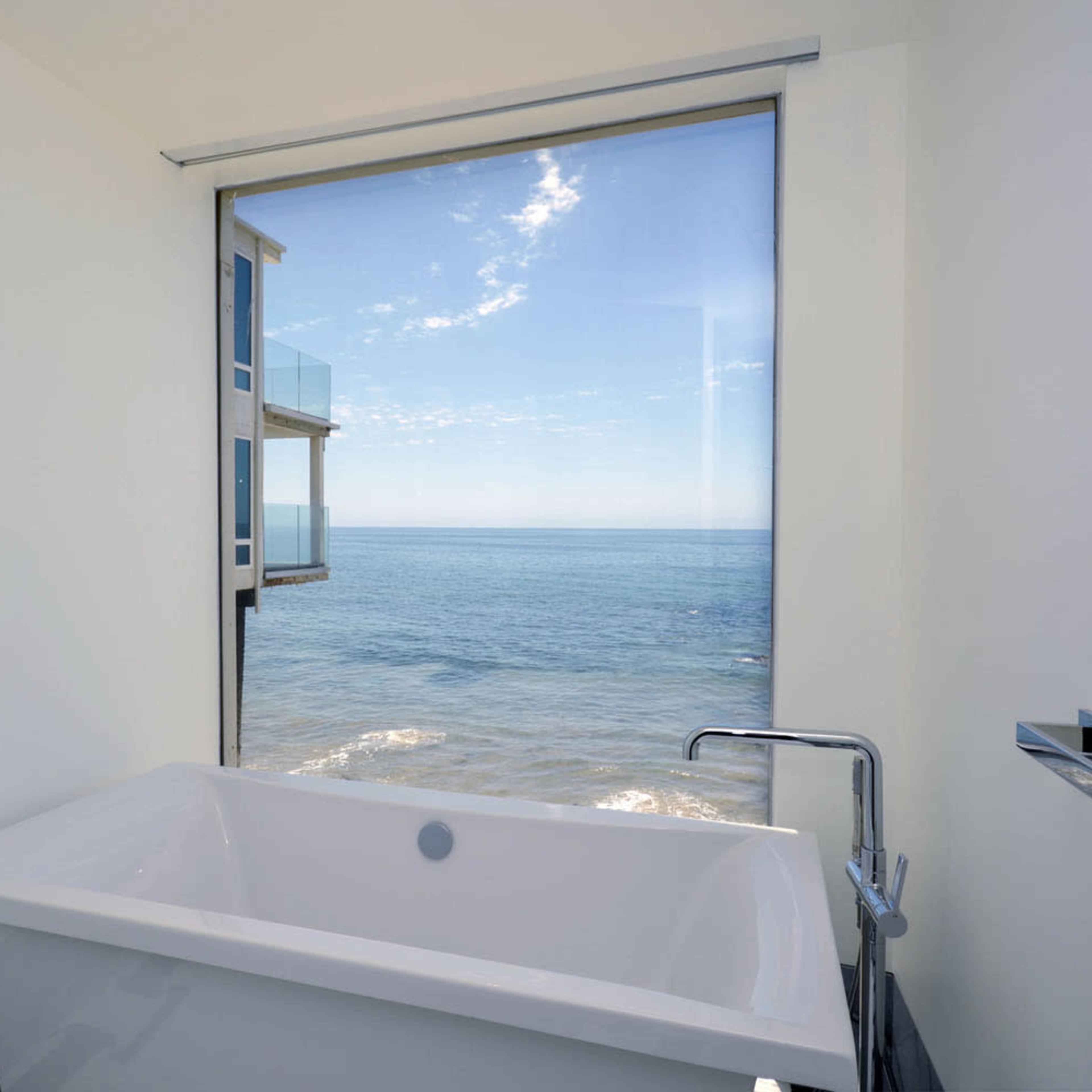 A modern bathtub sits in front of a large window overlooking the ocean and a clear sky.